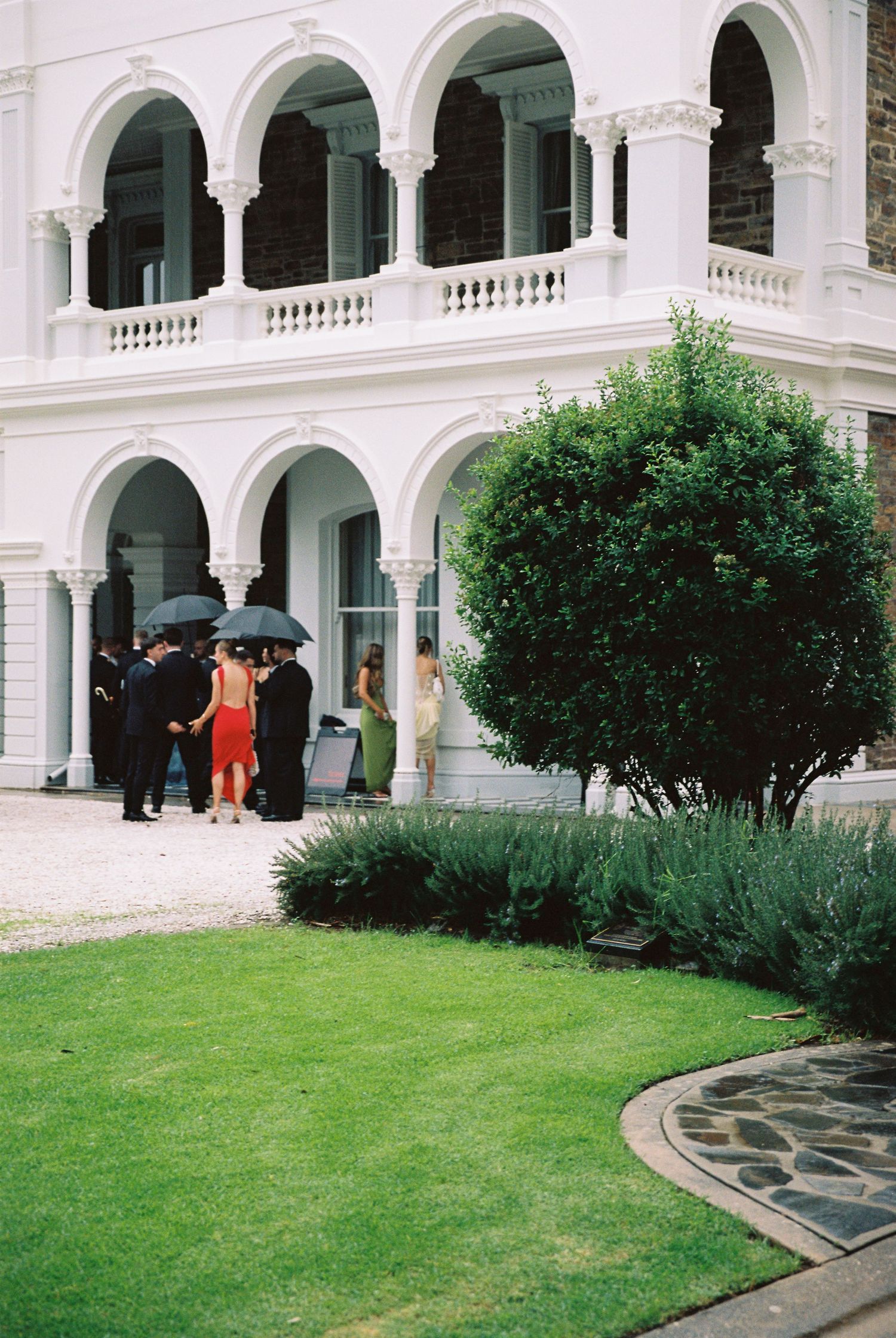 Wedding guests gathering under umbrellas outside elegant white colonial building with arched colonnade.