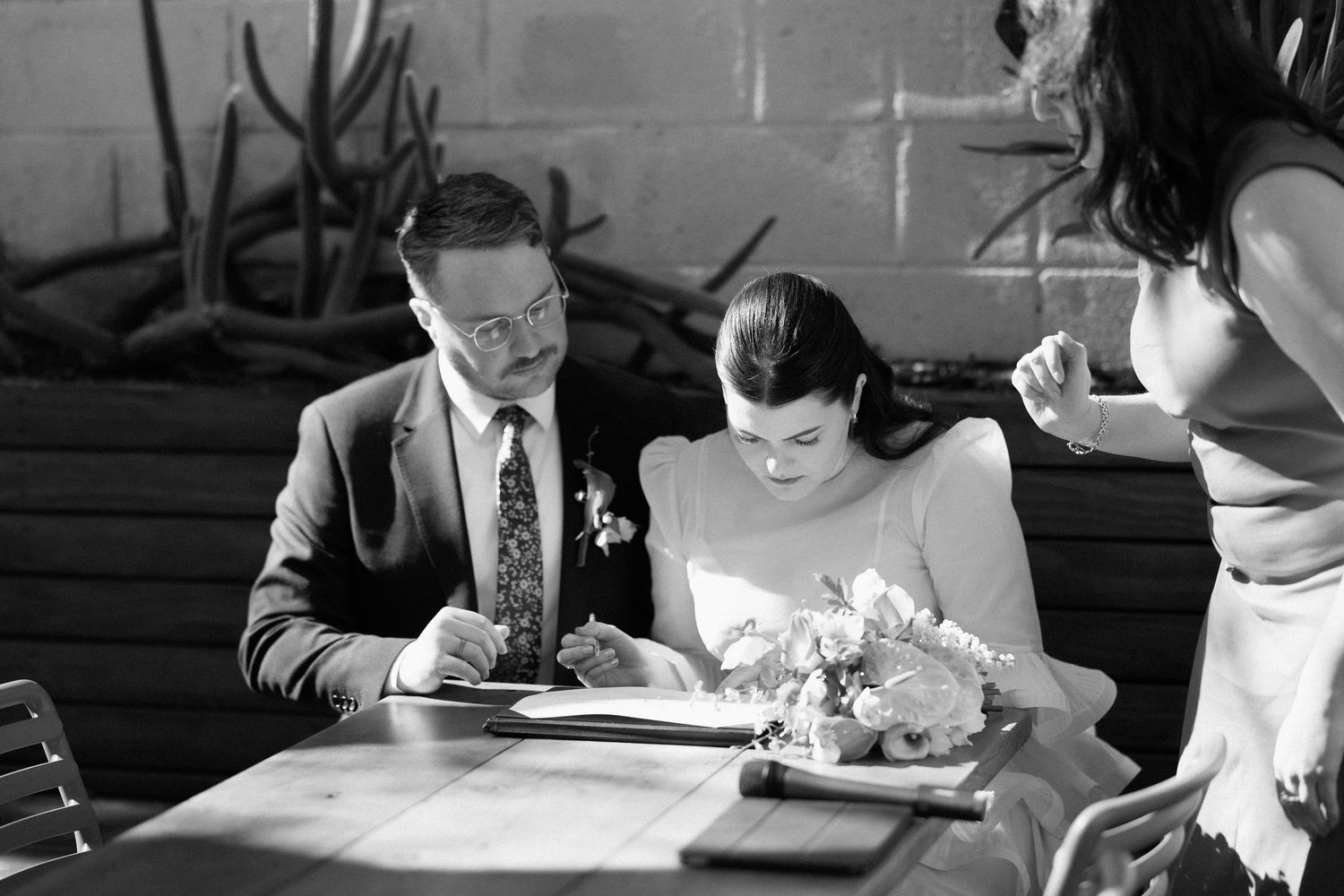 Black and white photo of bride and groom signing wedding register at a rustic wooden table.