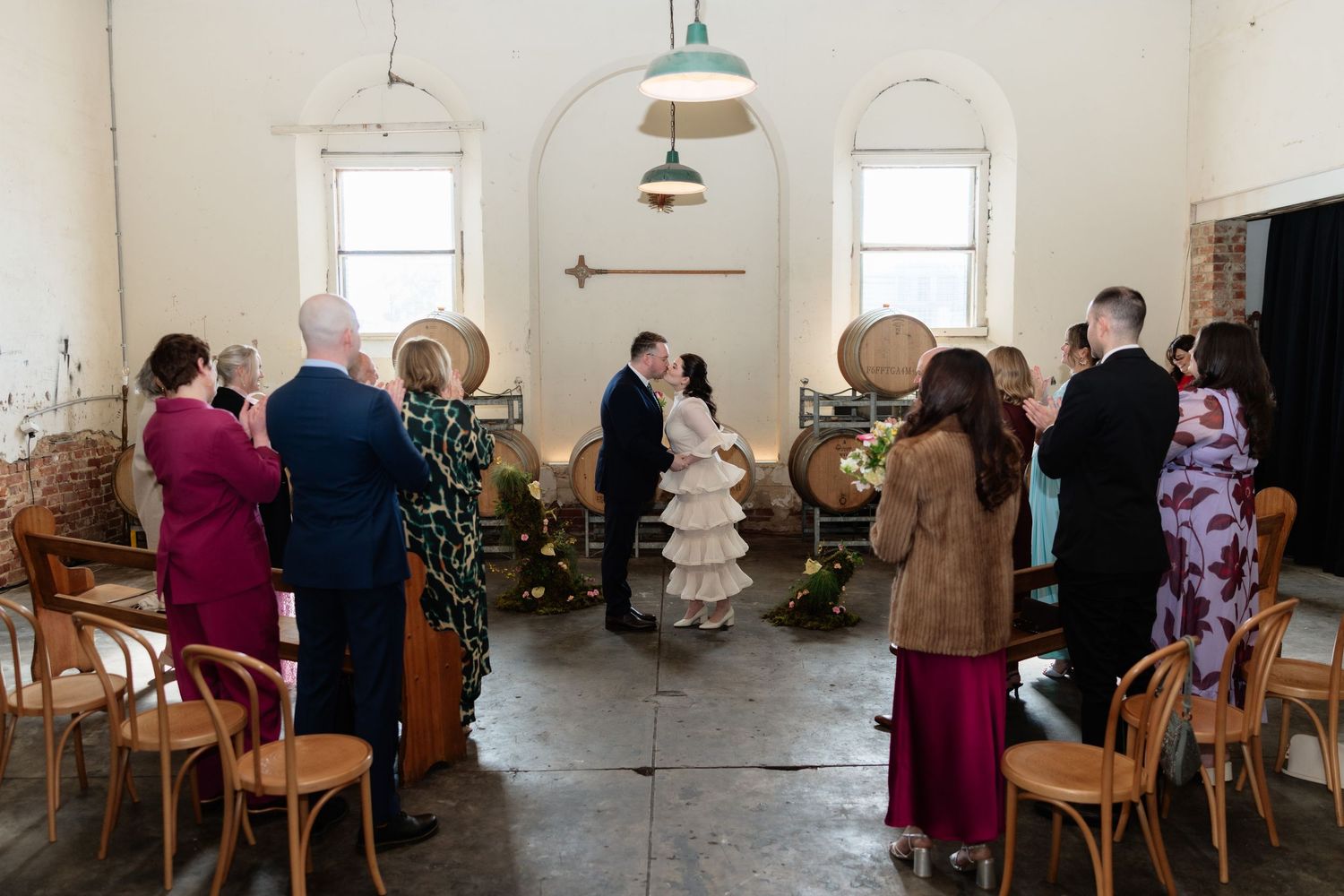 Intimate wedding ceremony in a rustic venue with wooden barrels, bentwood chairs, and guests standing as couple exchanges vows.