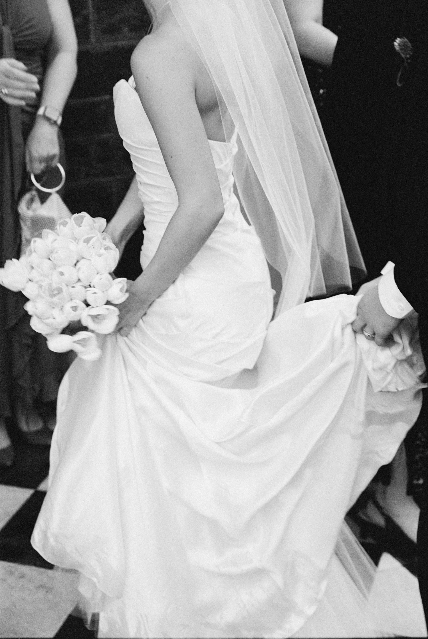 Black and white photo of a bride in a strapless gown holding a white bouquet, with veil flowing down her back.