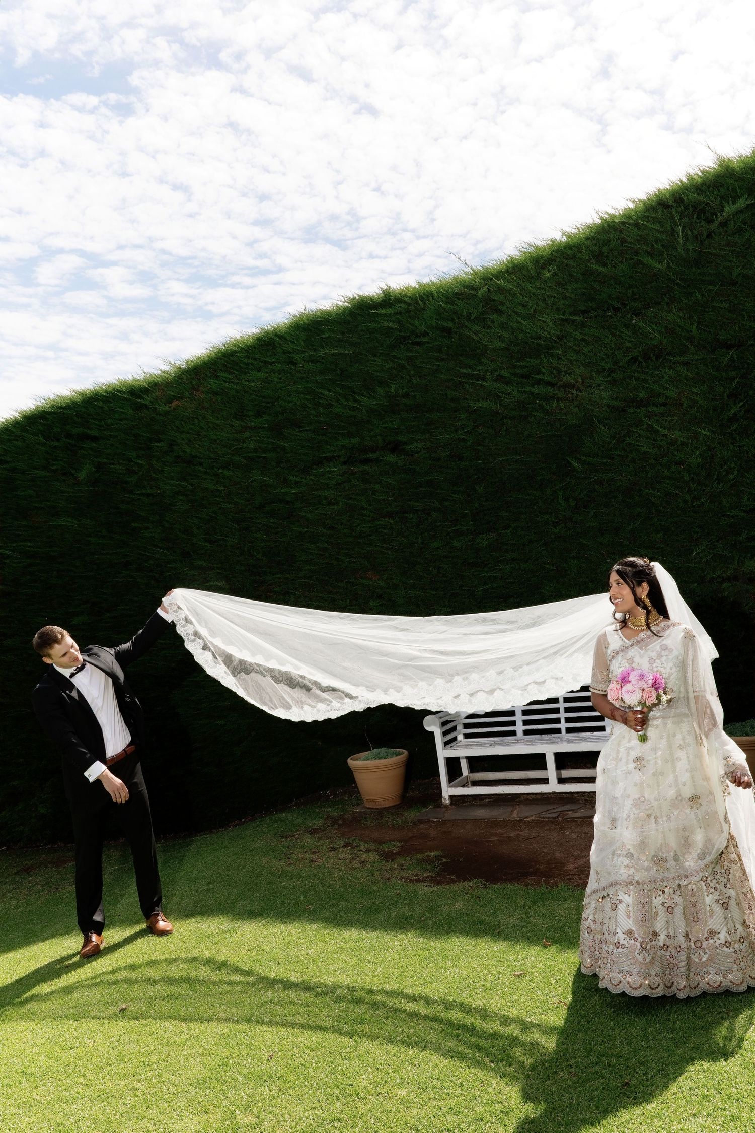 Groom in tuxedo holds flowing white bridal veil as bride in embroidered gown holds pink bouquet in garden.