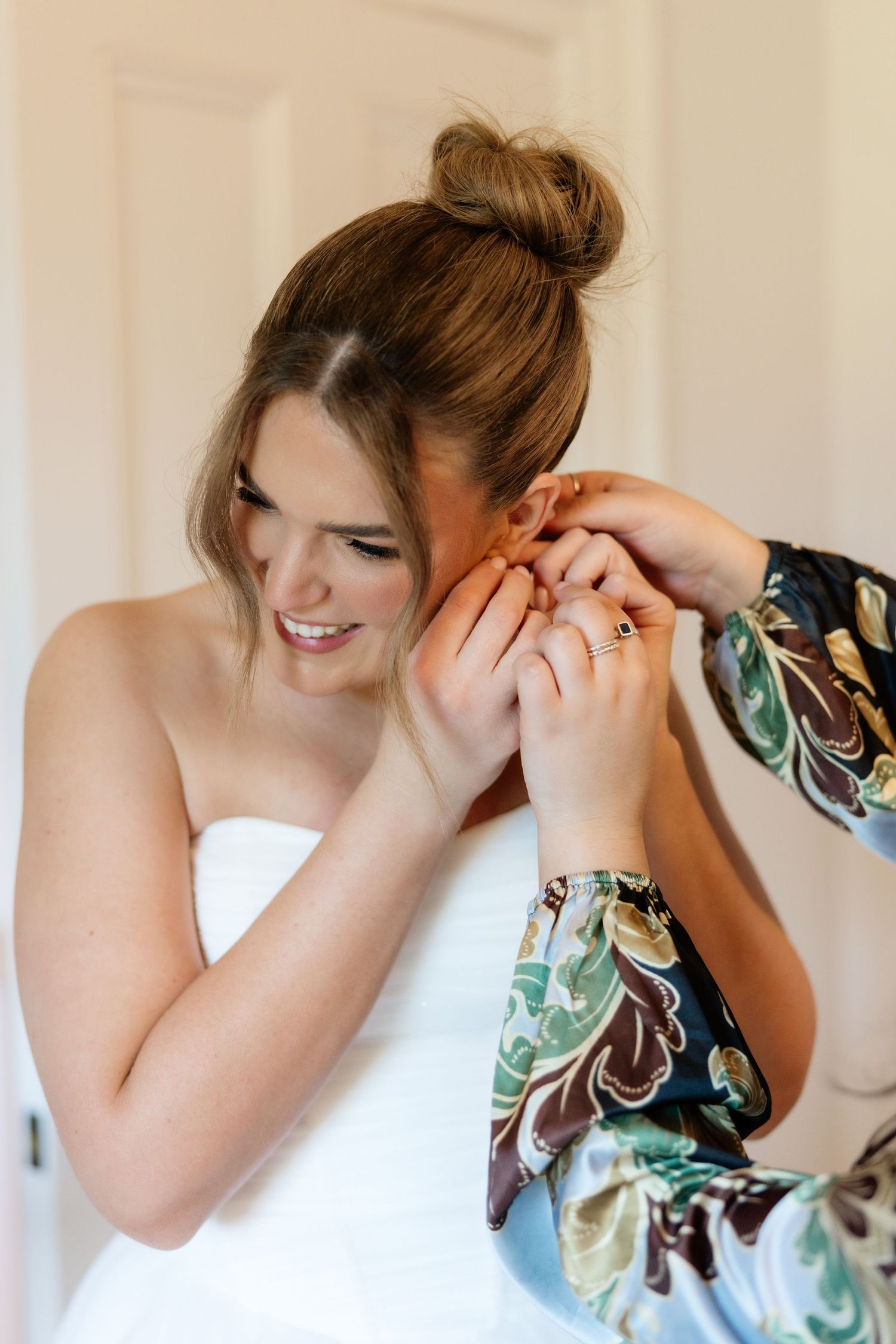 Bride in white strapless gown getting earring put on by bridesmaid in floral robe during wedding preparations.