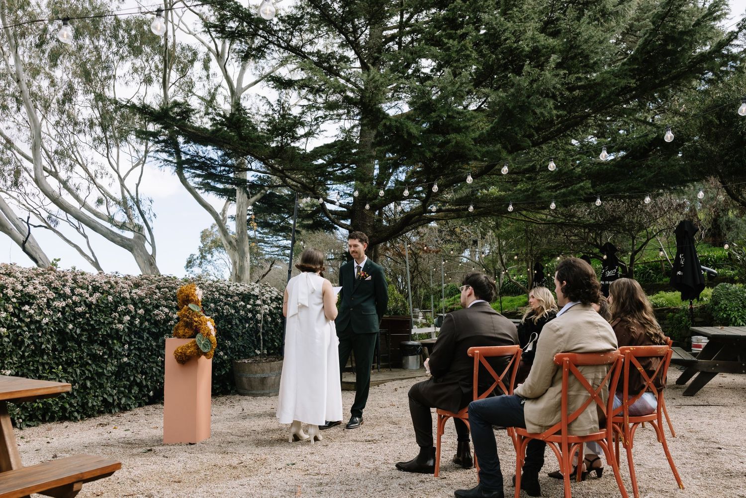Outdoor wedding ceremony with couple exchanging vows under trees, guests seated on orange chairs in a garden setting.
