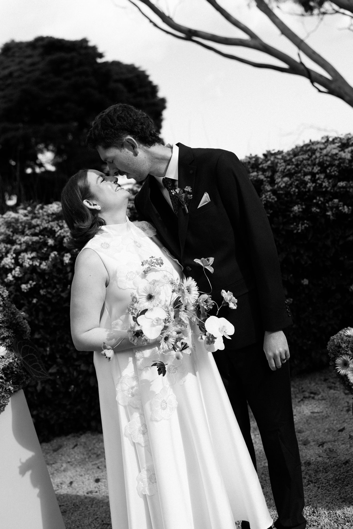 Black and white photo of a bride and groom sharing a kiss outdoors, bride holding a floral bouquet in a garden setting.