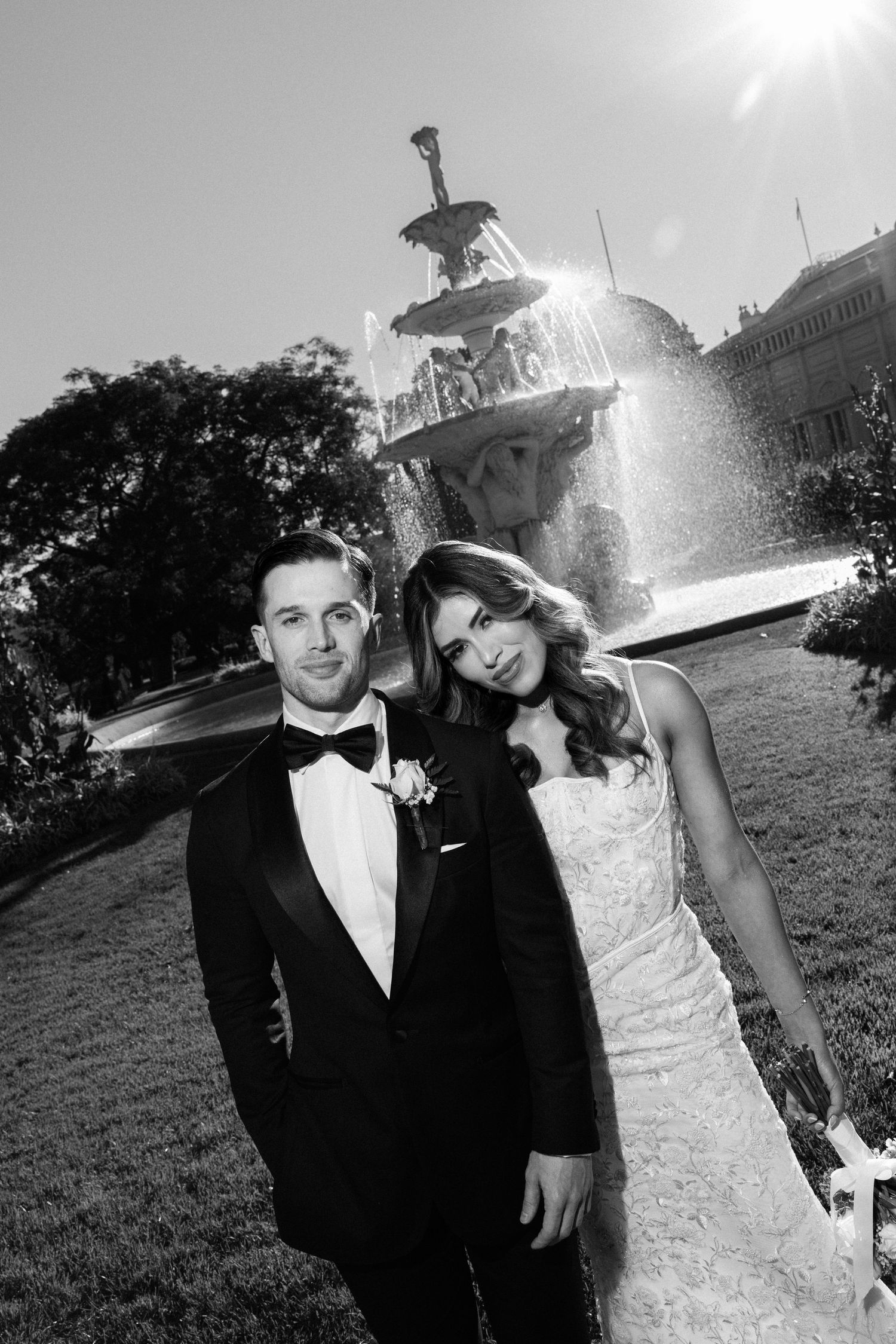 Black and white wedding photo of bride and groom posing in front of an ornate fountain in a garden.