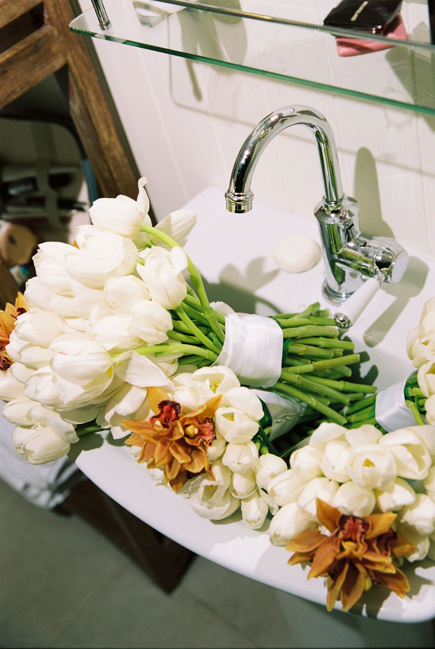 White tulips and orange orchids arranged in a white bathroom sink with a chrome faucet, creating an elegant floral display.