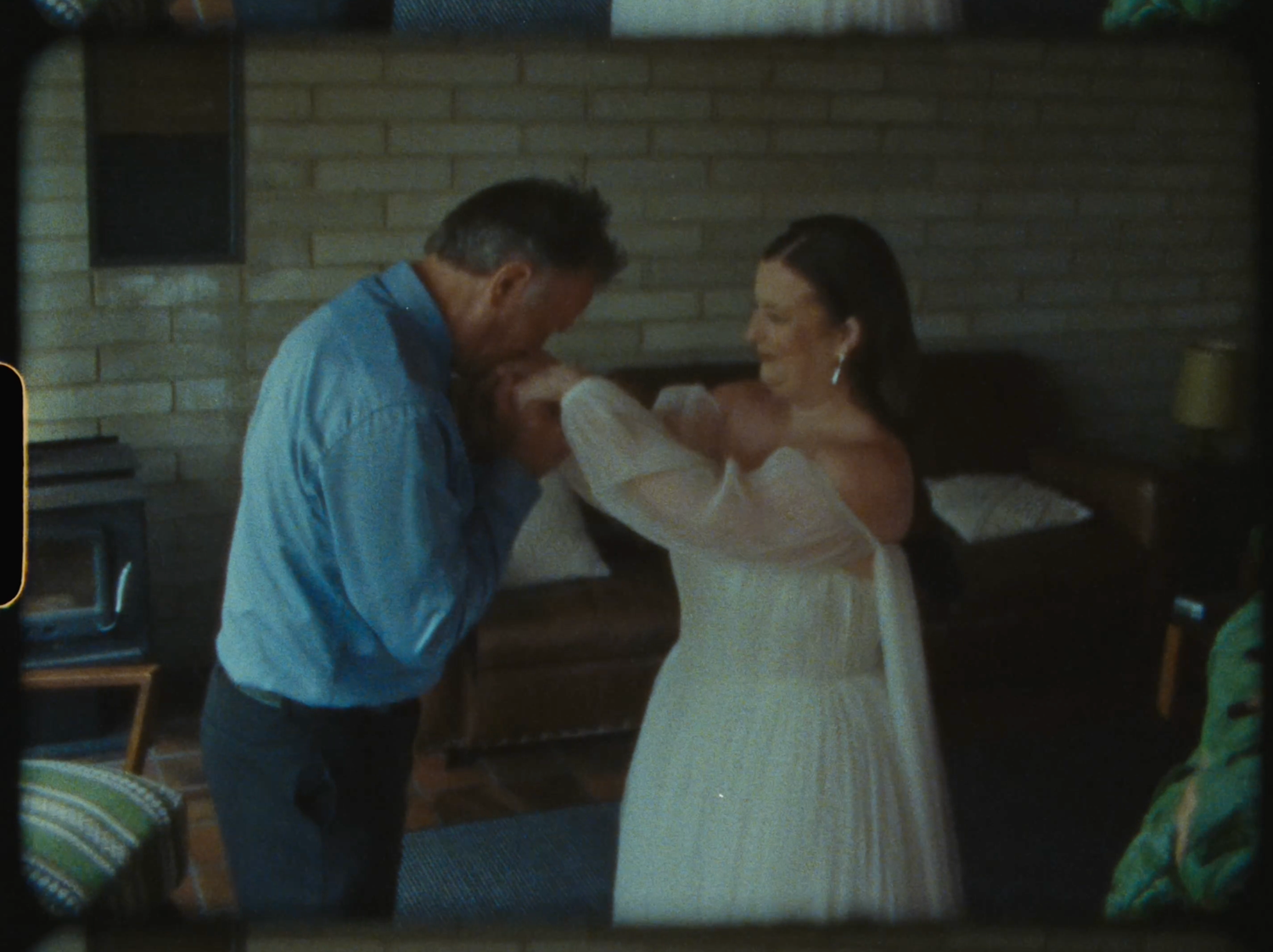 A man in a blue shirt kisses a woman's hand in a white dress indoors.
