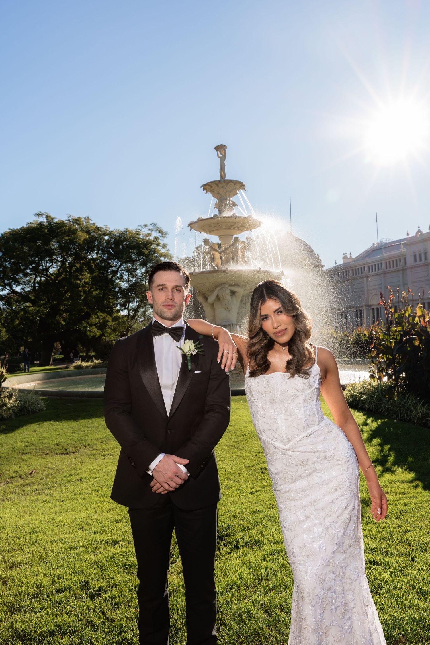 Bride and groom posing in front of a sunlit fountain in a lush garden on their wedding day.