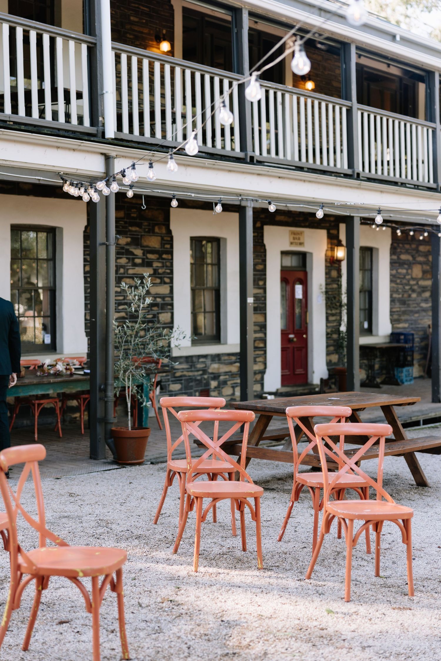 Rustic outdoor dining area with coral chairs and string lights outside a stone building with a red door.