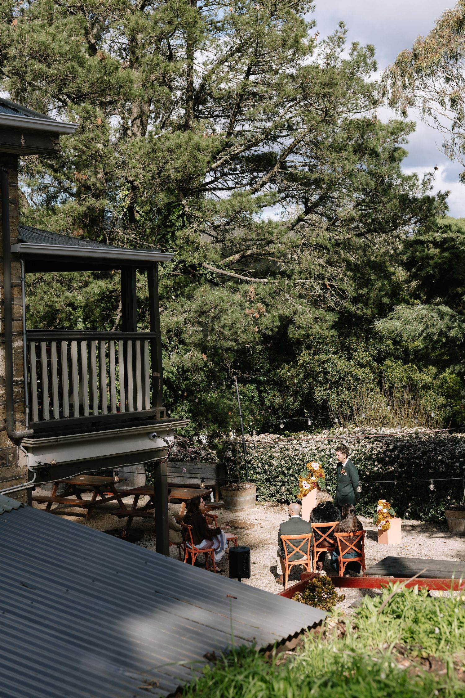 Rustic outdoor dining area with wooden deck, bistro chairs, and lush green trees in a charming garden setting.