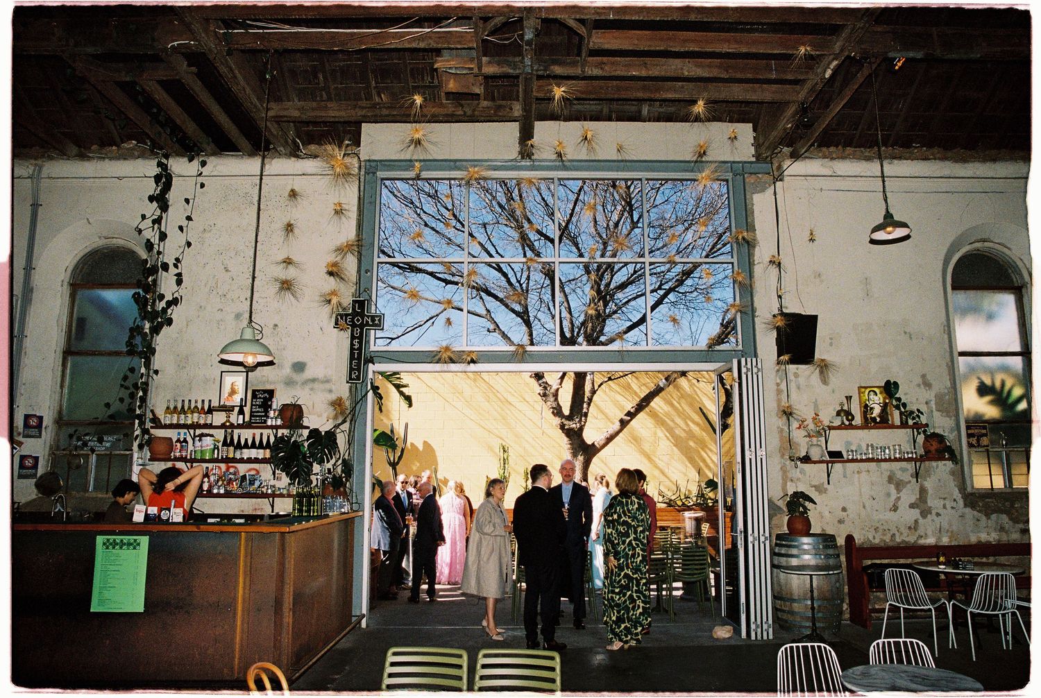 Rustic wedding venue interior with exposed brick walls, bar area, and guests gathered near large open doors revealing a bare winter tree.