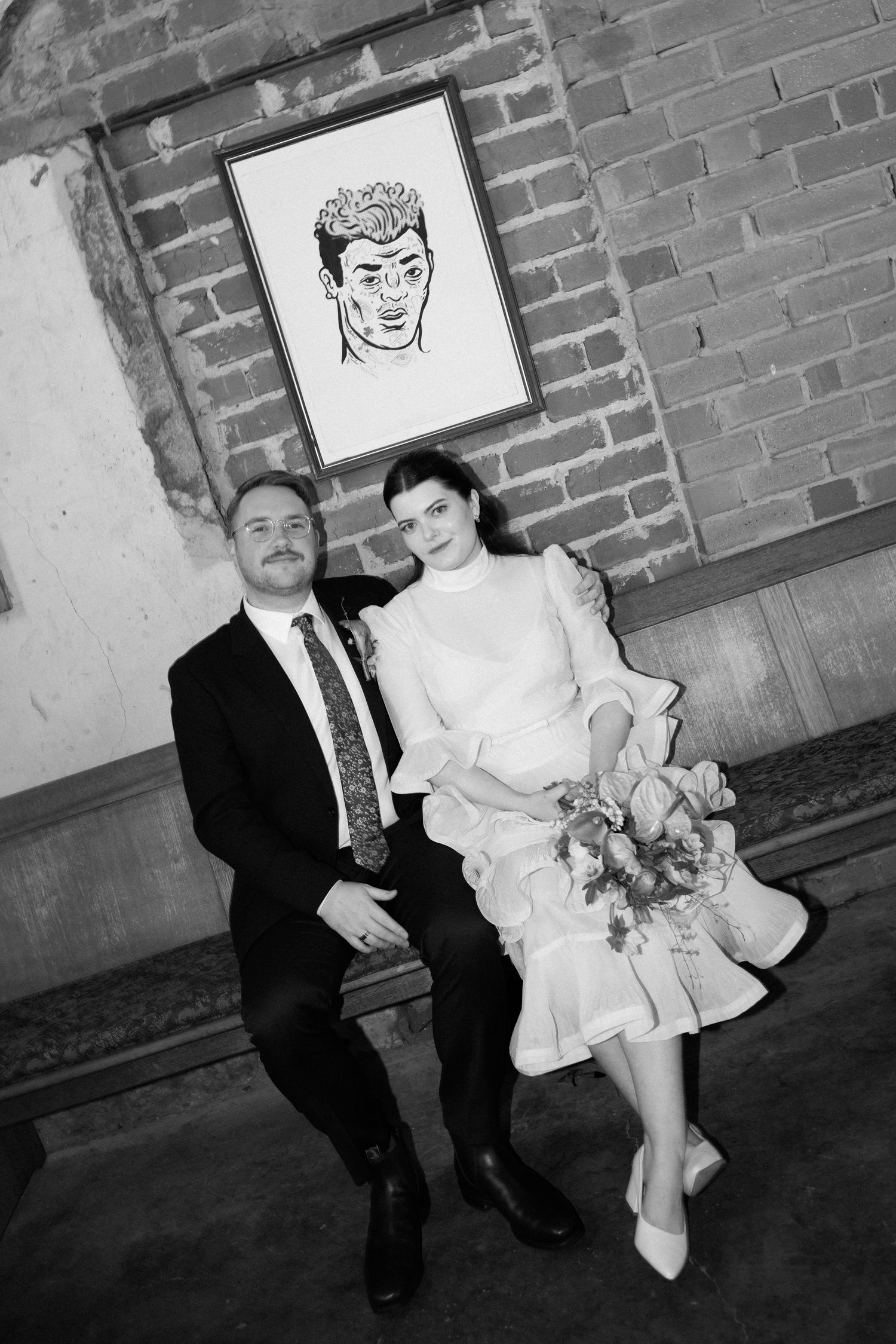 Black and white portrait of bride and groom seated together in front of brick wall artwork.