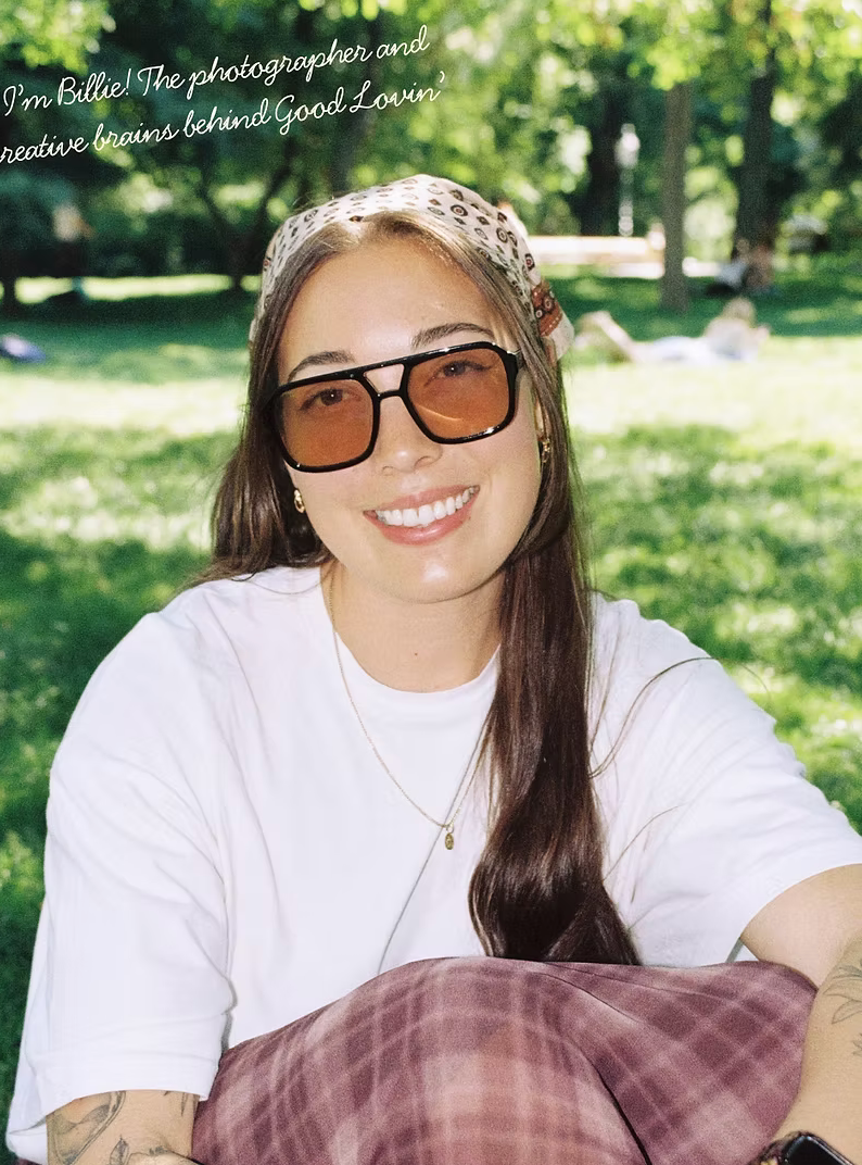 Smiling young woman with sunglasses and headband sitting outdoors in a sunny park wearing a white t-shirt.