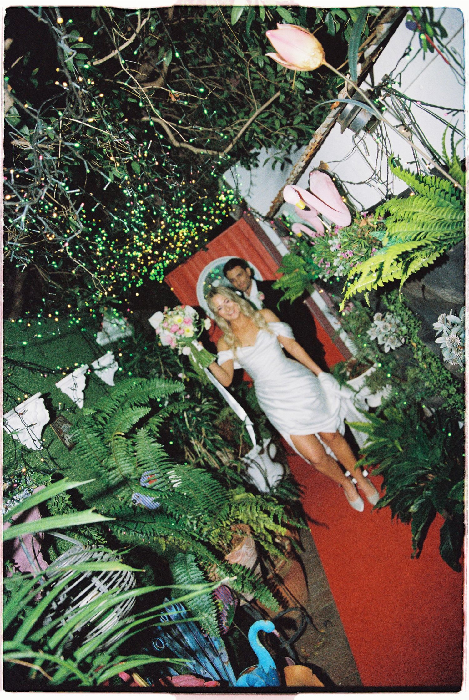 Bride in white mini dress holding bouquet poses with groom among lush tropical greenery and fairy lights at night.