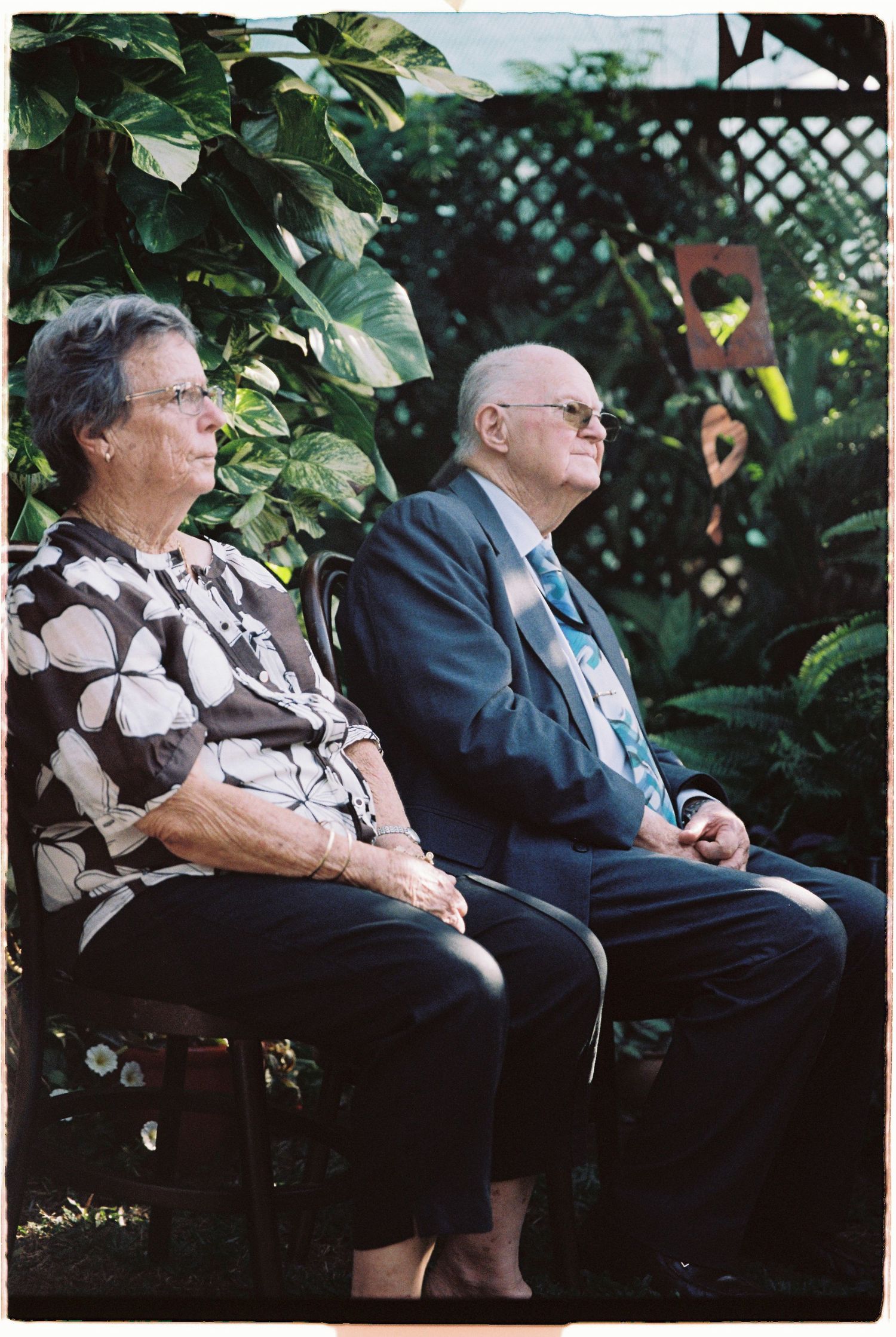 Elderly couple seated outdoors at a garden wedding ceremony surrounded by tropical plants.