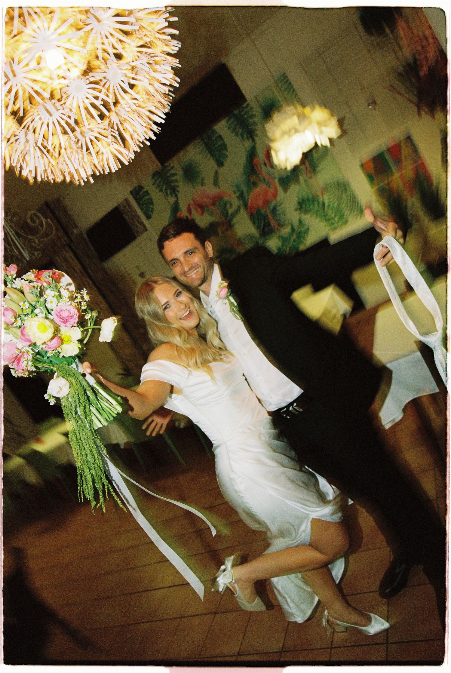 A joyful bride and groom pose playfully with a floral bouquet inside a decorated wedding venue.