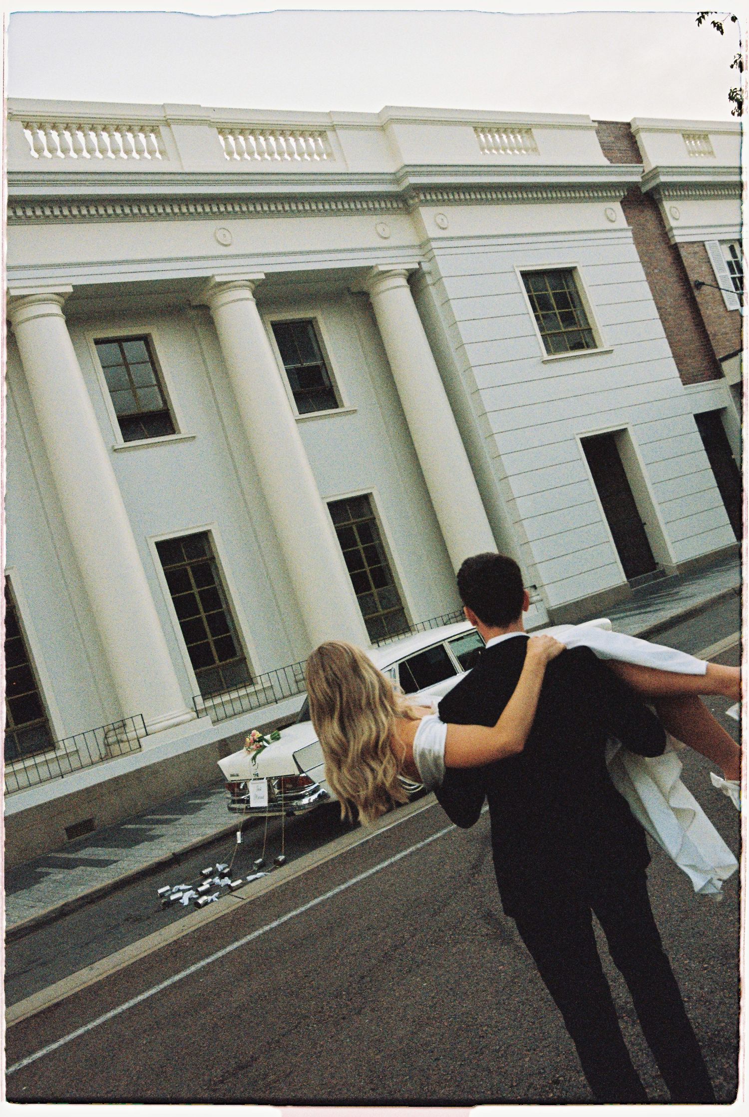 Groom carries bride in his arms toward a grand white neoclassical building on their wedding day.