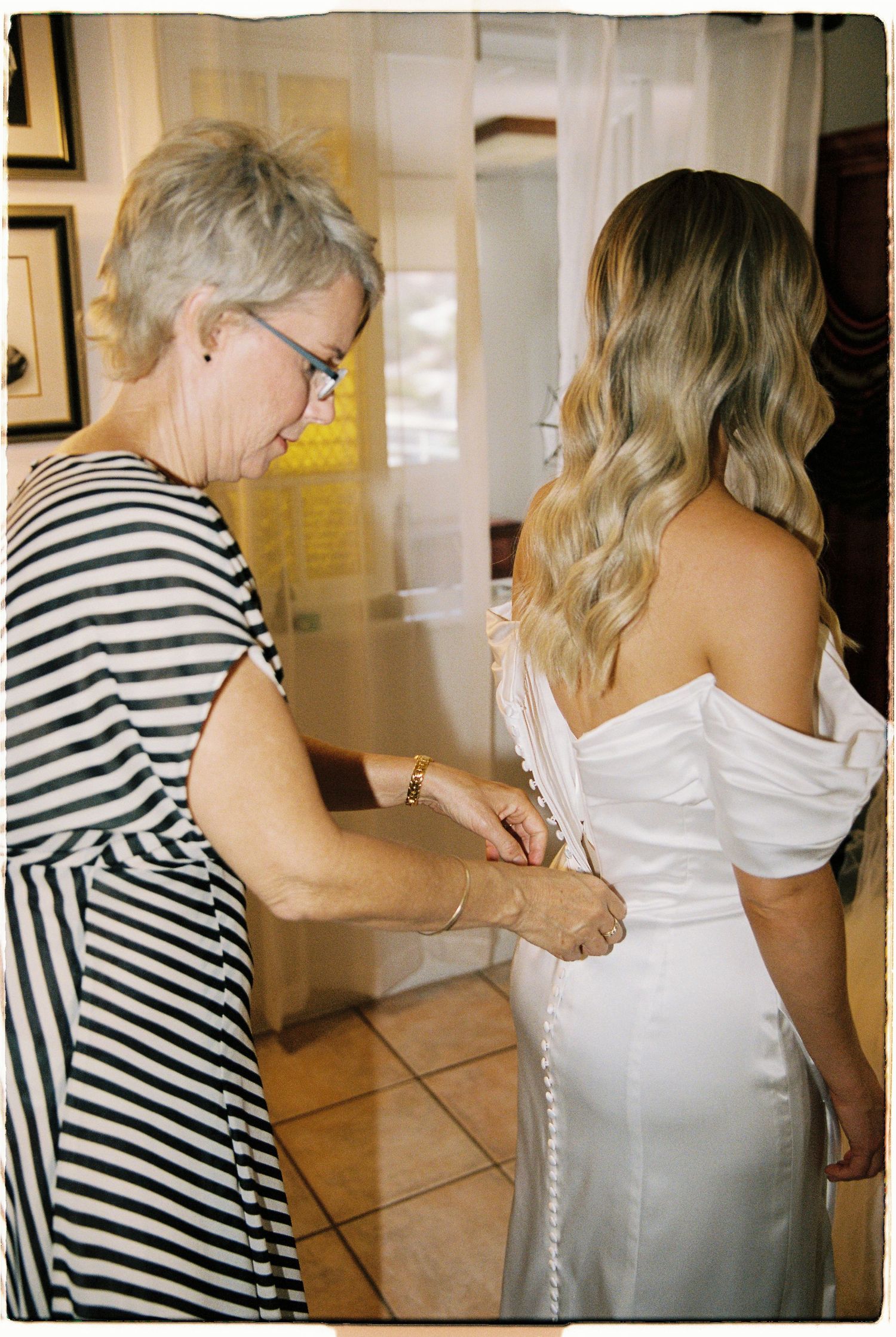 Older woman helping bride button up white off-shoulder wedding gown from behind in a warmly lit room.