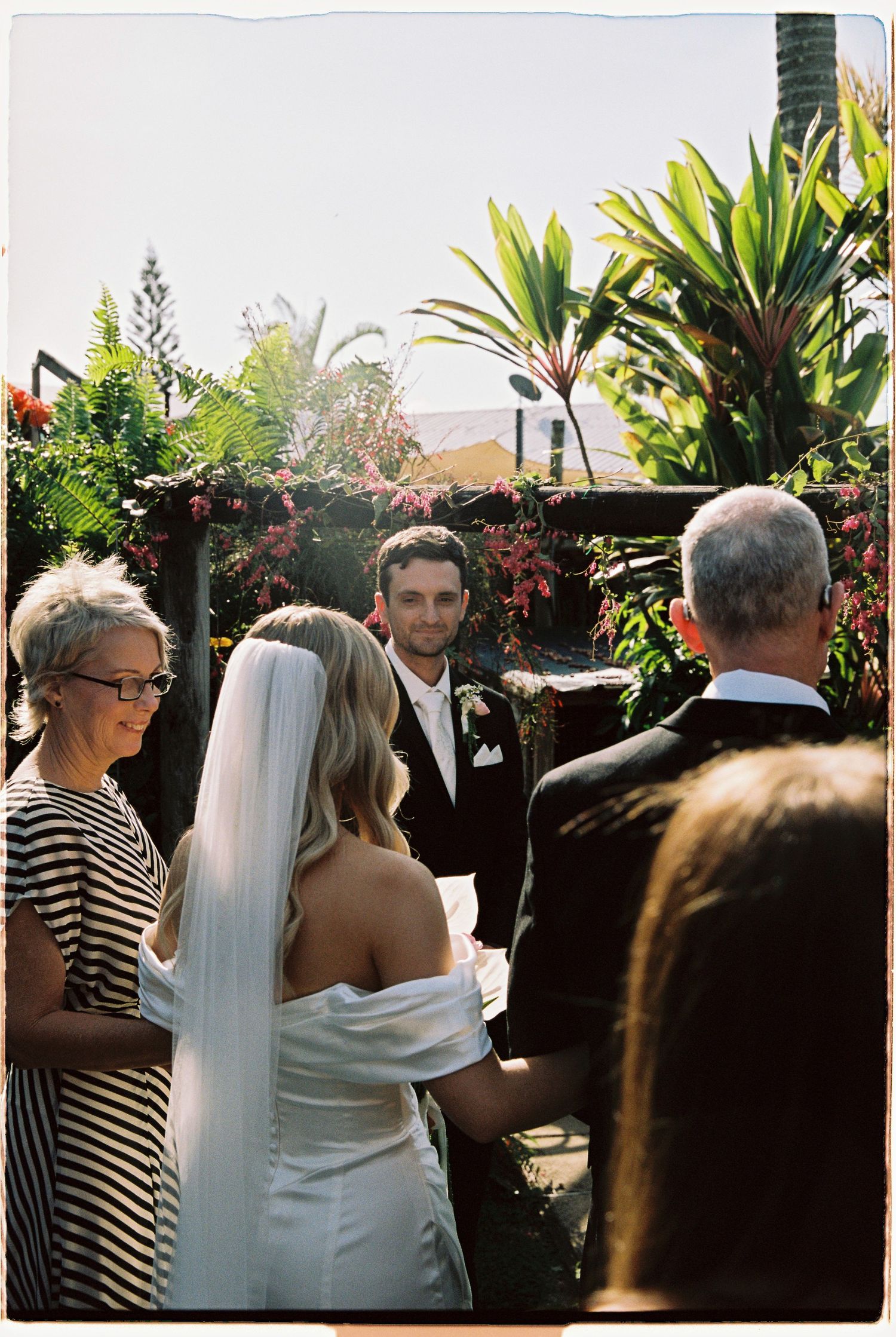 Groom in black tuxedo gazes at bride in white off-shoulder gown during outdoor garden wedding ceremony.