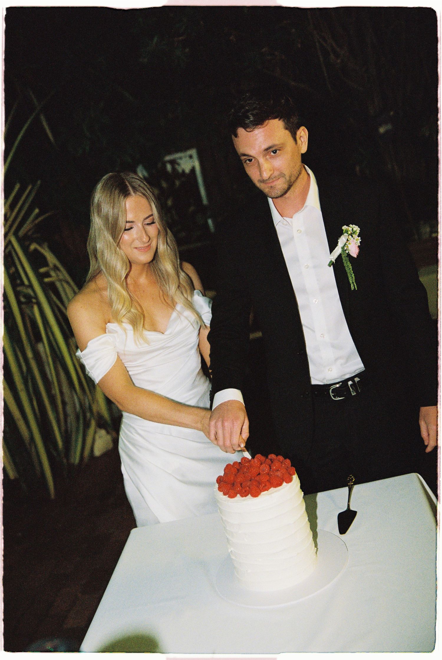Bride and groom cutting white wedding cake topped with fresh red raspberries.