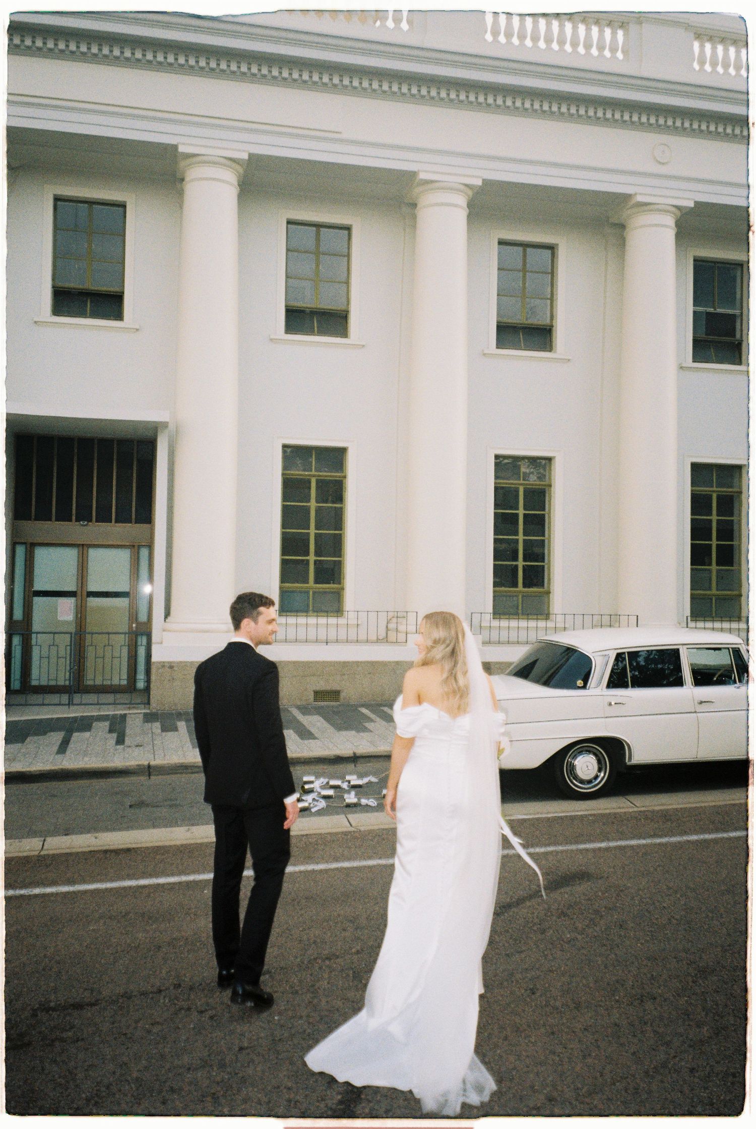 Elegant couple in wedding attire pose in front of a grand white columned building with a classic car.