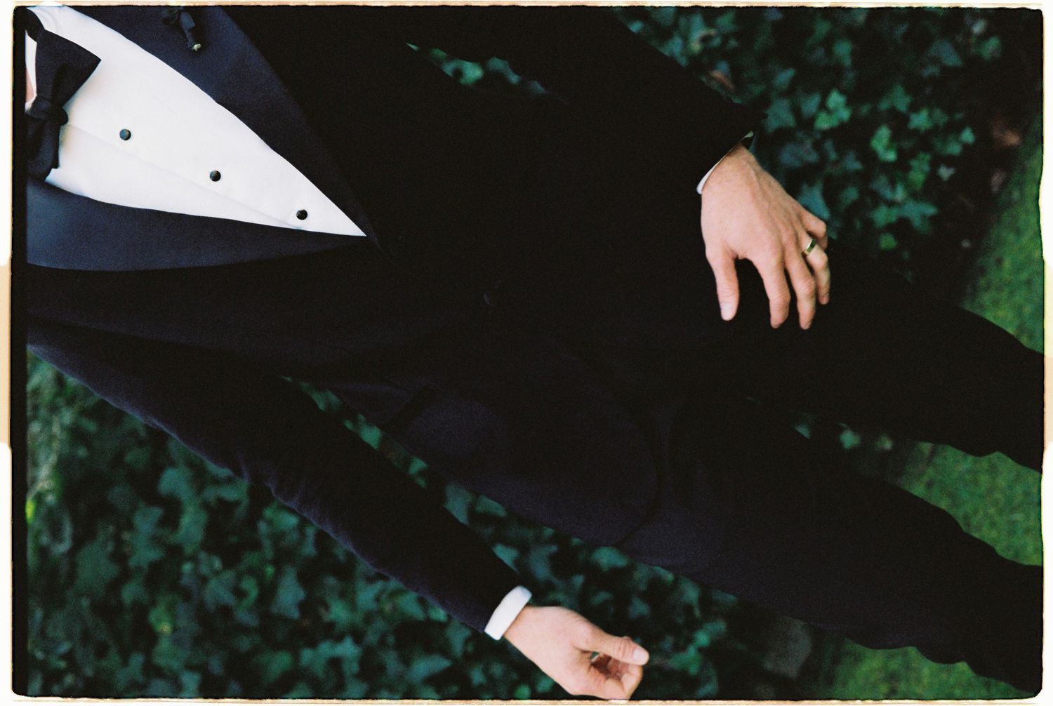 Aerial view of two grooms in black tuxedos standing on green grass, one wearing a gold ring.