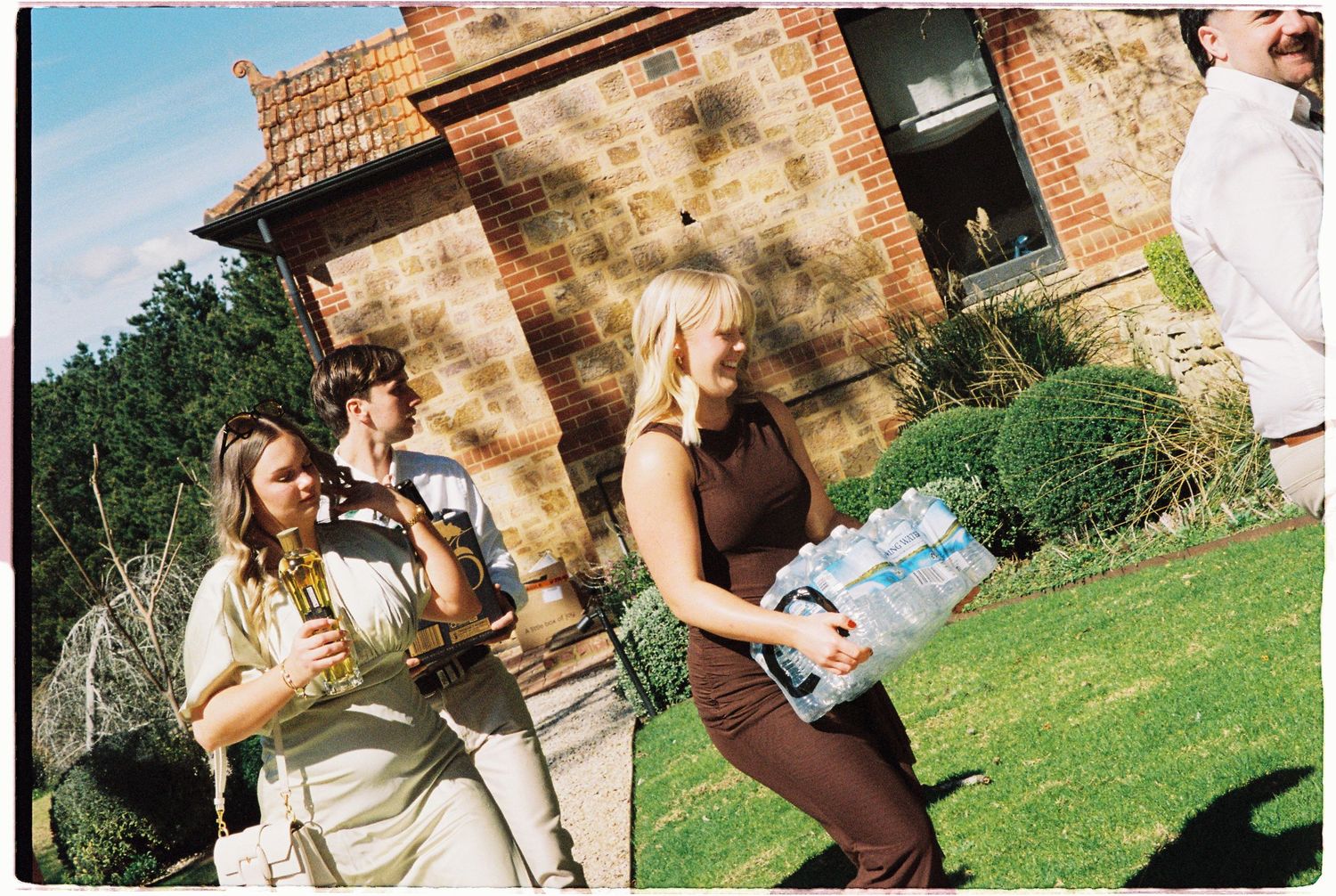 Wedding guests carrying supplies outside a rustic brick building on a sunny day.