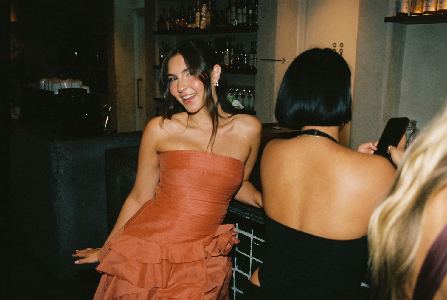 Woman in rust strapless dress smiles at wedding reception bar area.