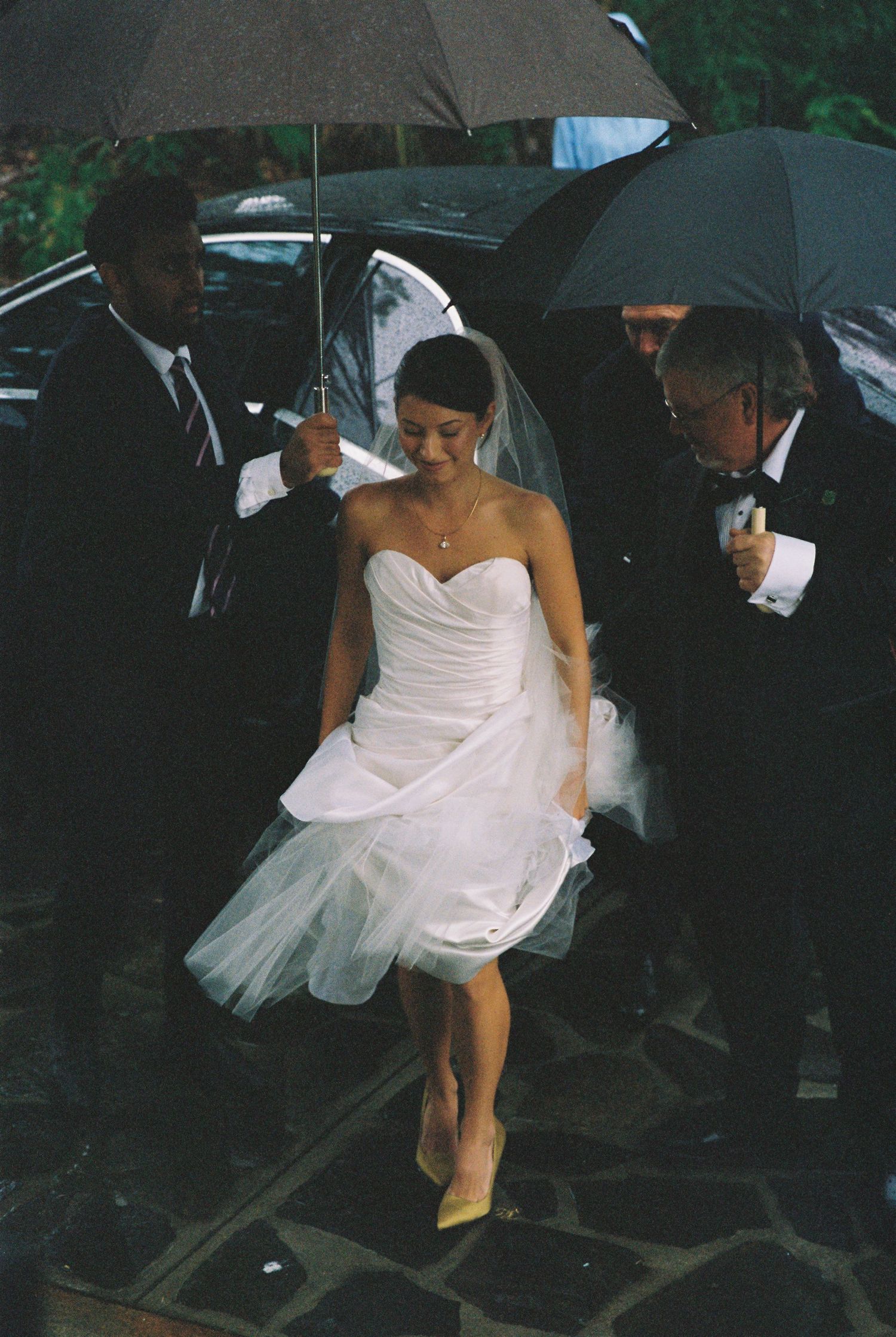 Bride in short white wedding dress with veil walks in rain escorted by guests holding black umbrellas.