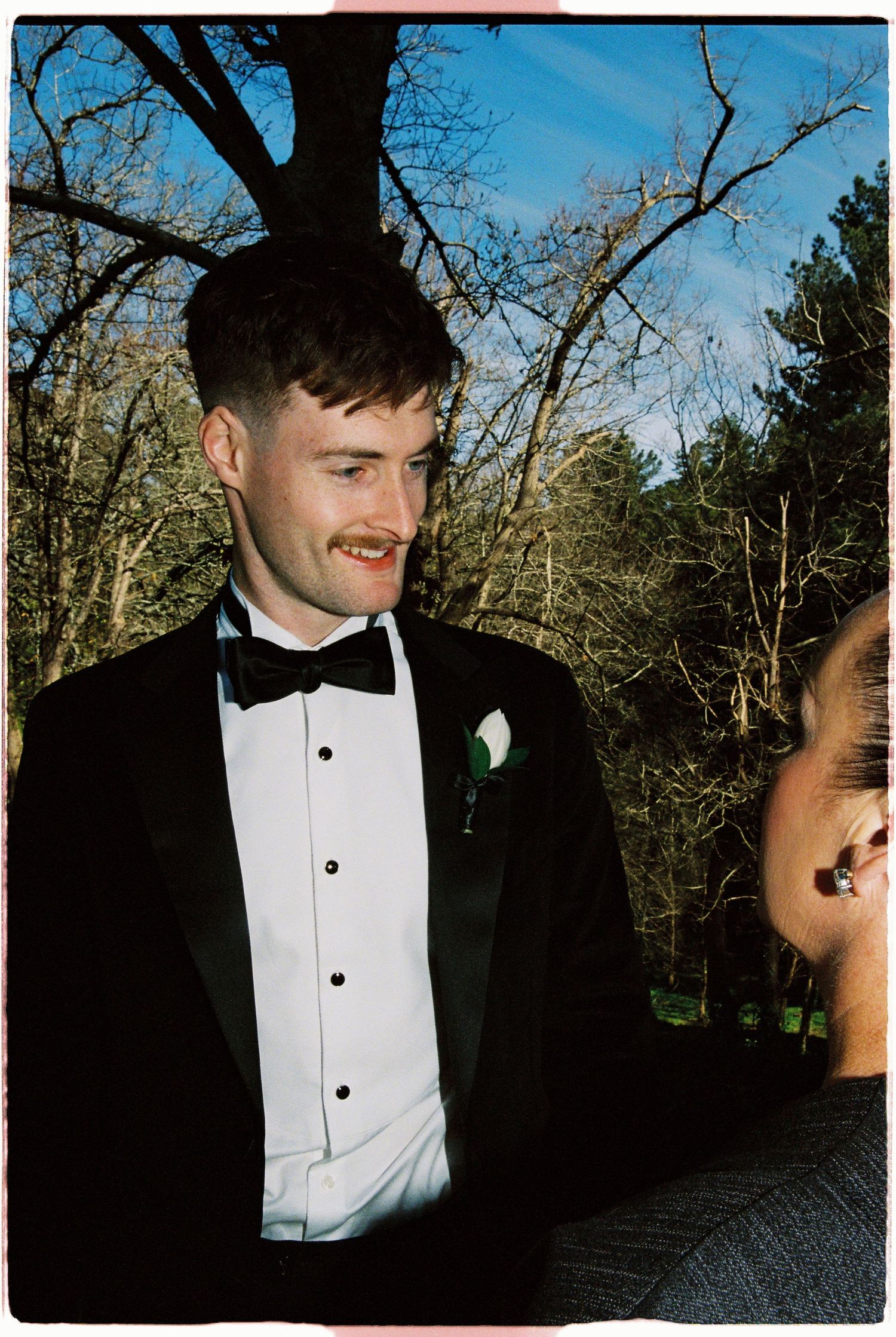 Groom in black tuxedo with bow tie and boutonniere smiling outdoors at a wedding ceremony.