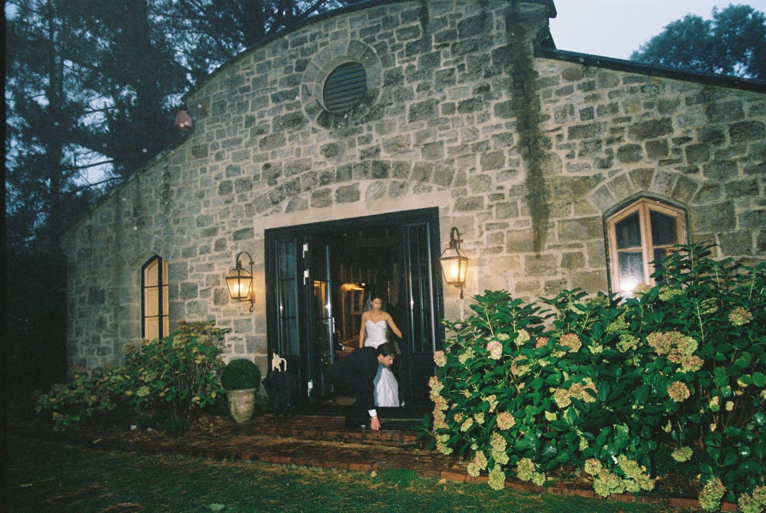 Stone building wedding venue entrance glows with warm lights surrounded by lush greenery at dusk.