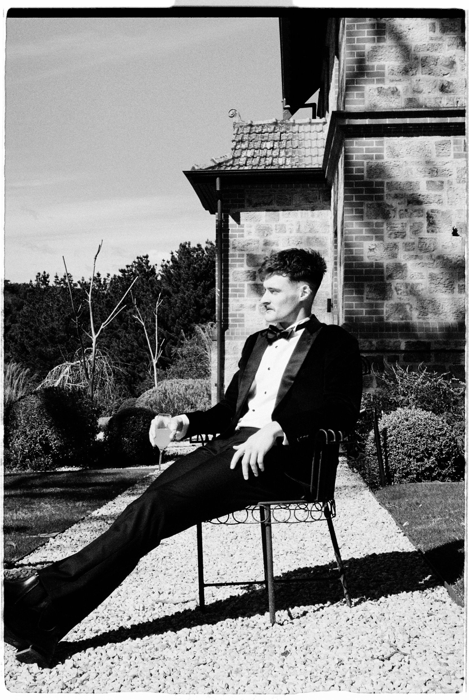 Groom in tuxedo relaxing on chair outside brick building in black and white wedding photo.