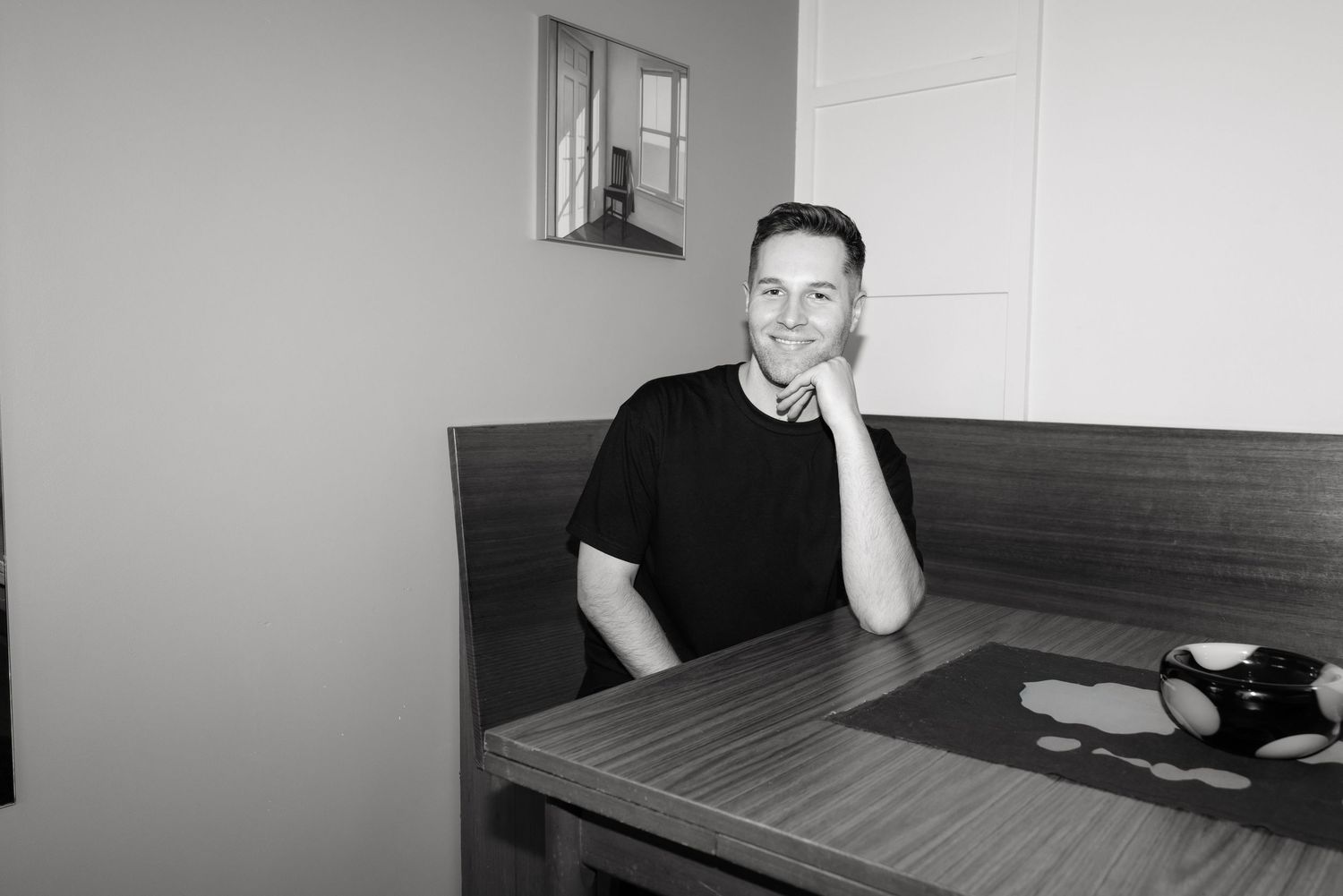 Black and white photo of a smiling man sitting at a wooden table in a cozy dining nook, resting his chin on his hand.