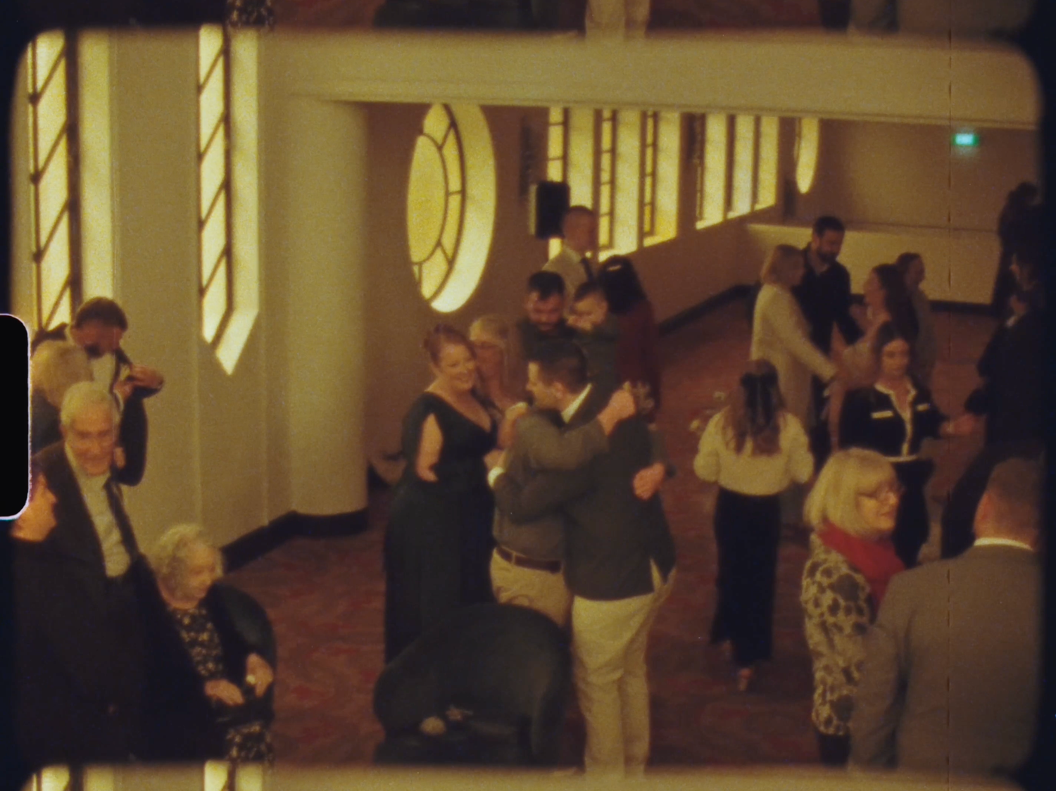Guests dancing and socializing in an elegant art deco ballroom with tall windows.