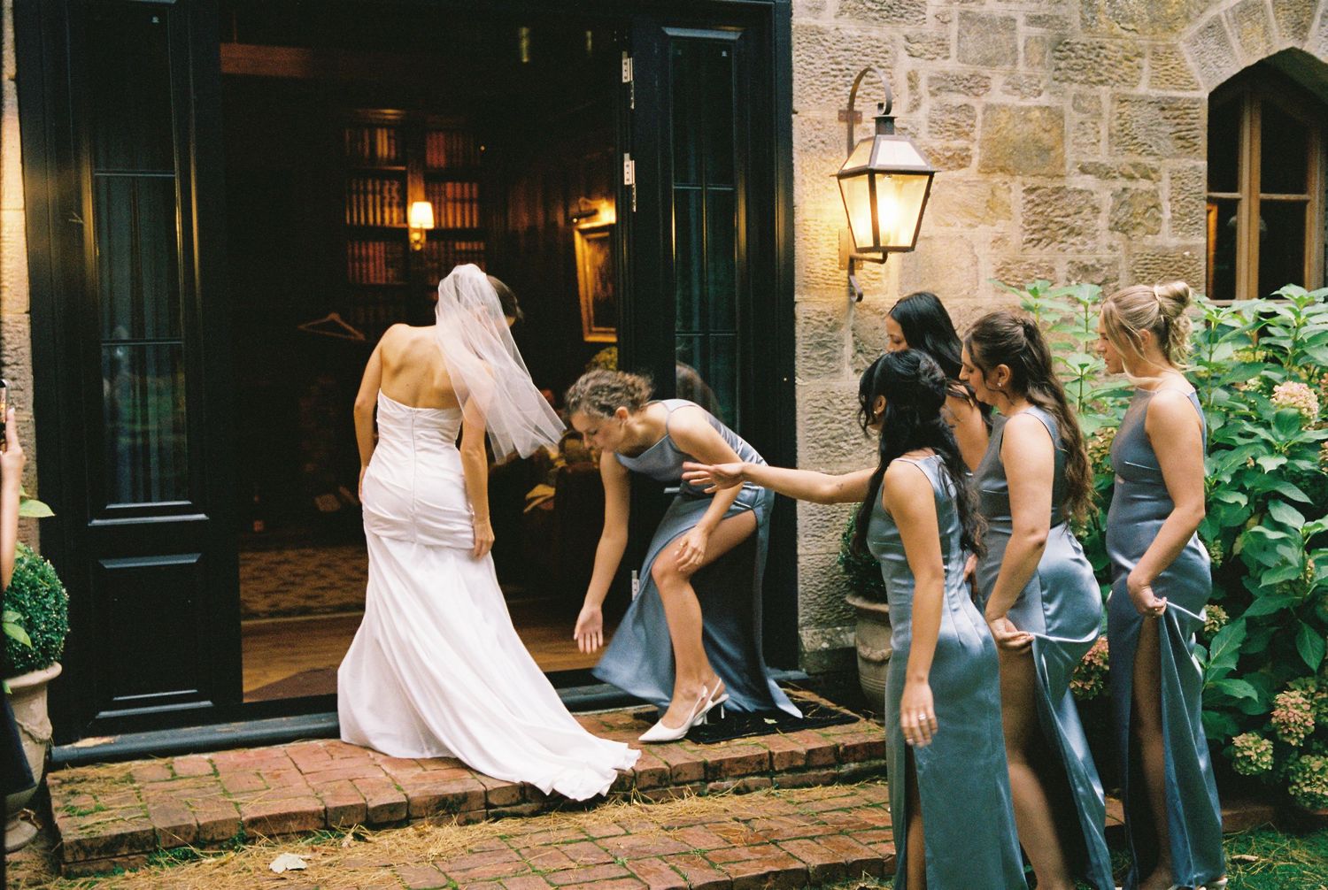 Bridesmaids in blue-grey gowns helping bride with wedding dress at stone venue entrance with lantern.