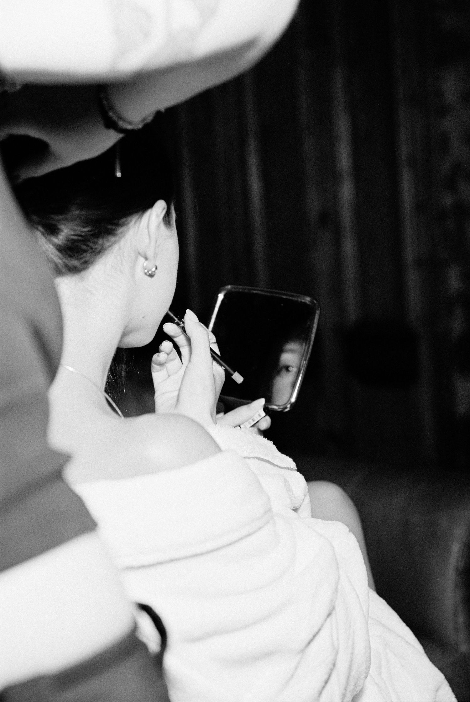 Bride applying earring using small handheld mirror in black and white wedding preparation photo.