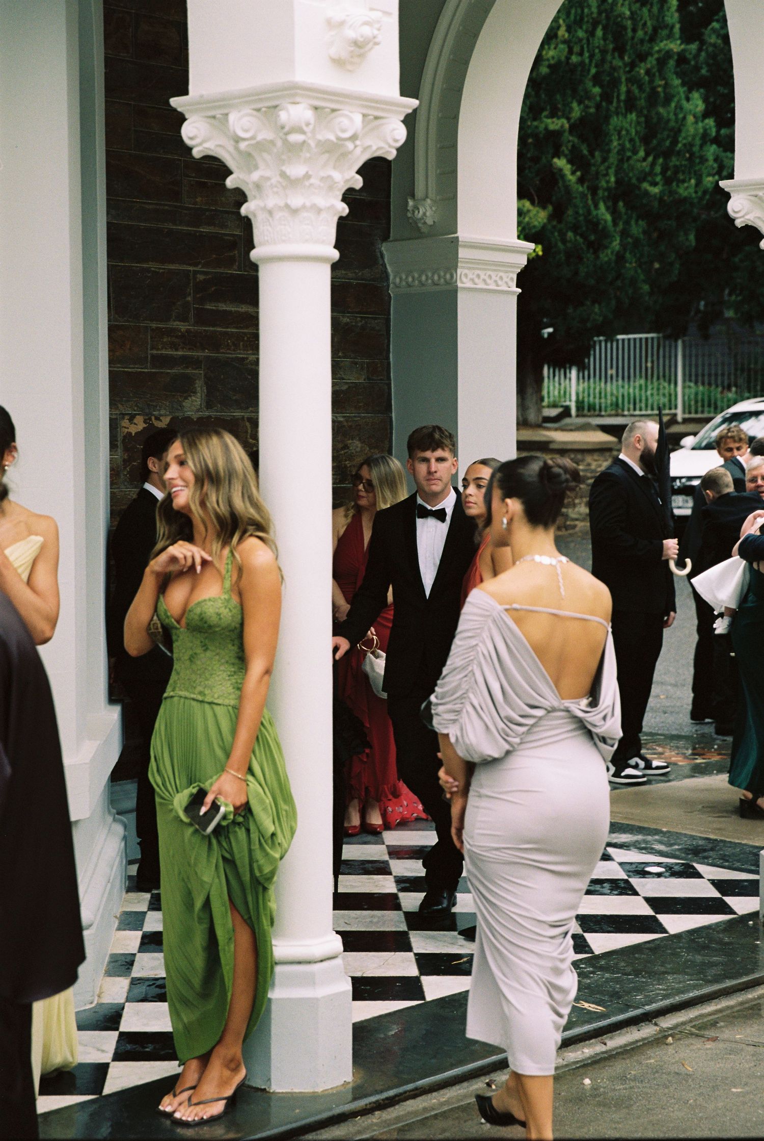 Wedding guests in formal attire socializing on a black and white checkered floor beneath ornate white columns.