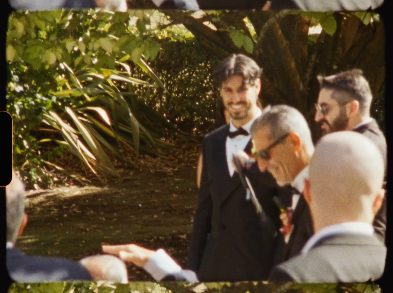Groom in black tuxedo smiling joyfully with wedding guests in a sunlit garden setting.