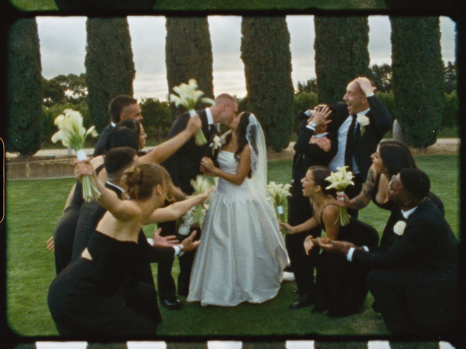 Joyful wedding party surrounding kissing bride and groom on lawn with cypress trees at golden hour.