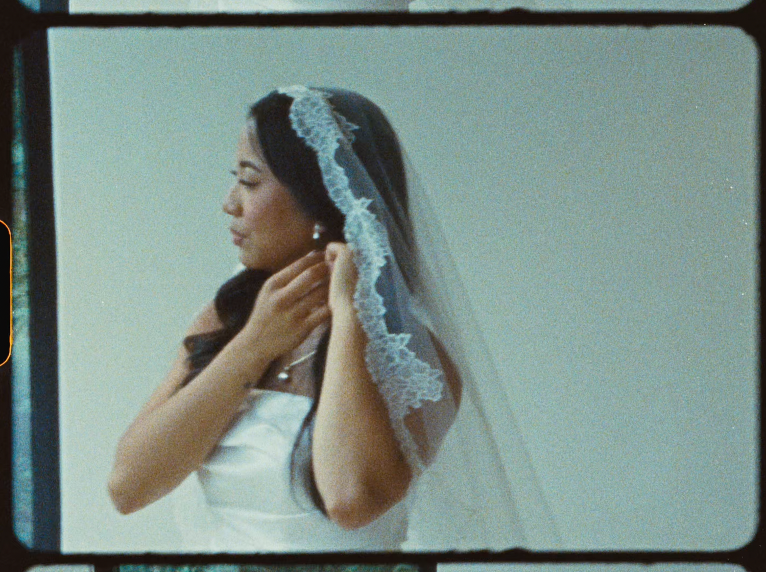 Bride in a white gown and lace-trimmed veil adjusts her earring while looking to the side.
