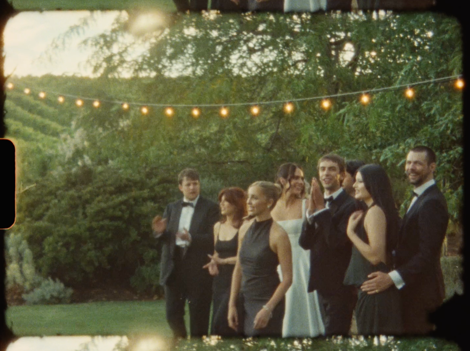 Wedding party poses under string lights in a lush garden at golden hour.