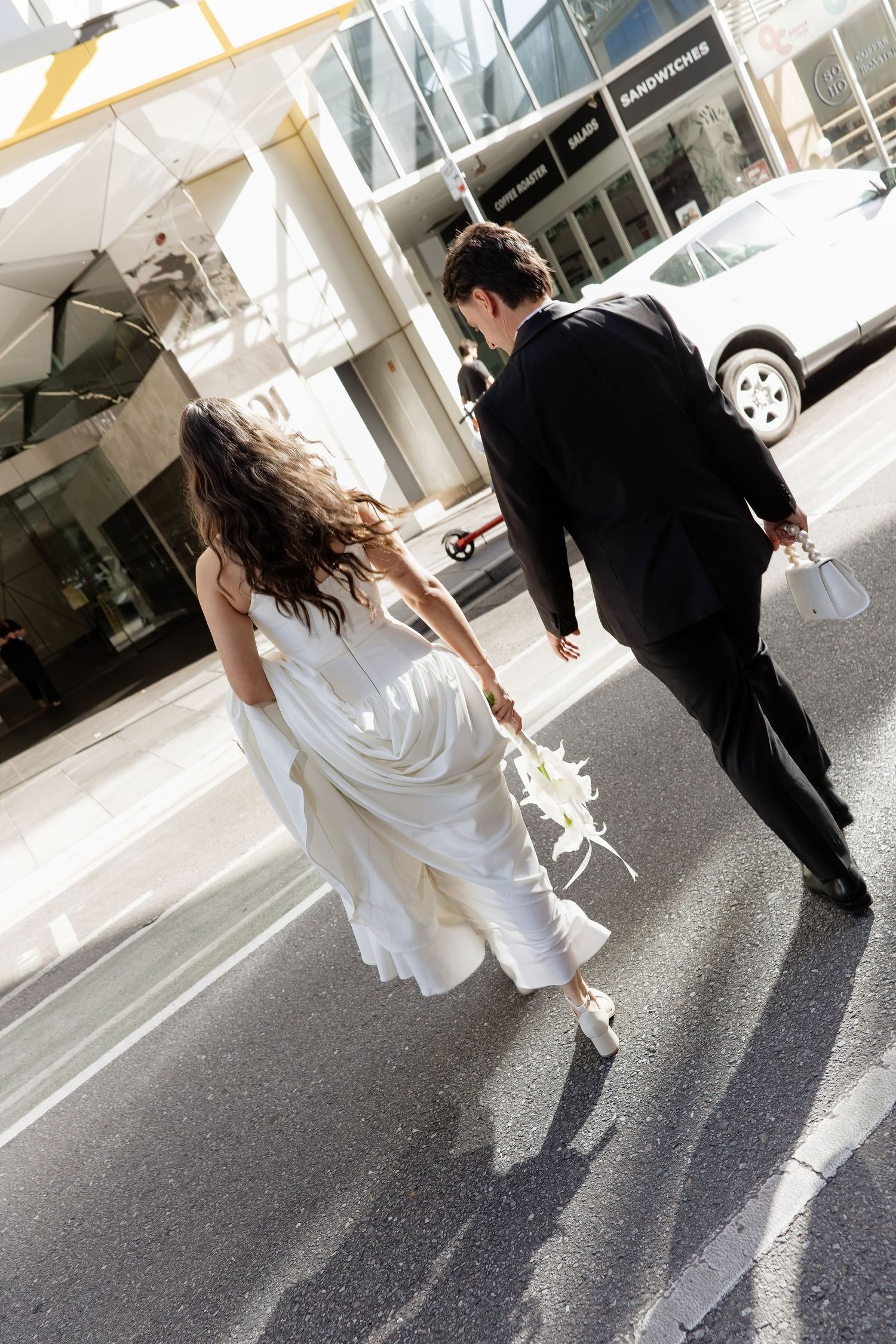 Bride in white dress and groom in black suit walking across city street holding bouquet of flowers.