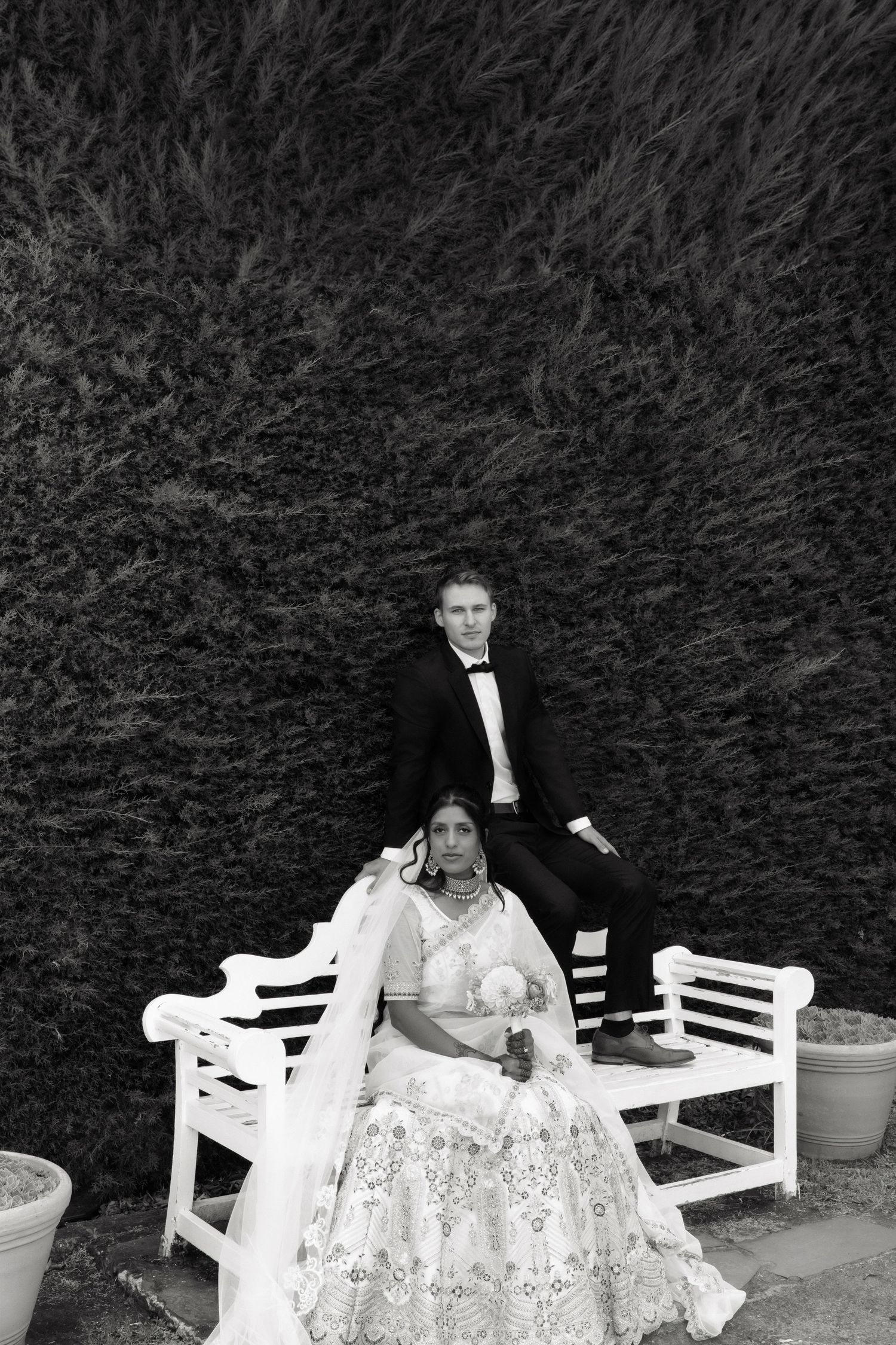 Bride in embroidered lehenga and groom in tuxedo pose on white bench against dark hedge.