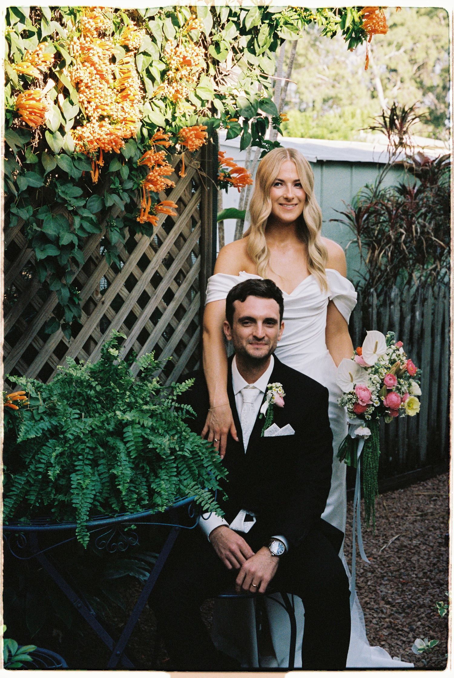 Bride standing behind seated groom in garden with vibrant orange flowering vines at wedding.