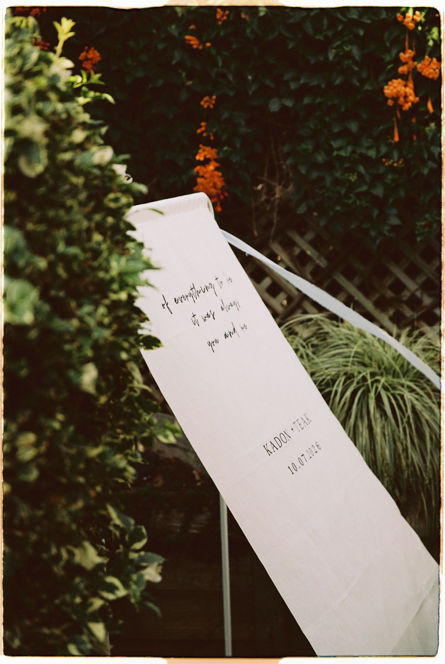 White wedding vow scroll with handwritten text displayed outdoors among lush green garden hedges.