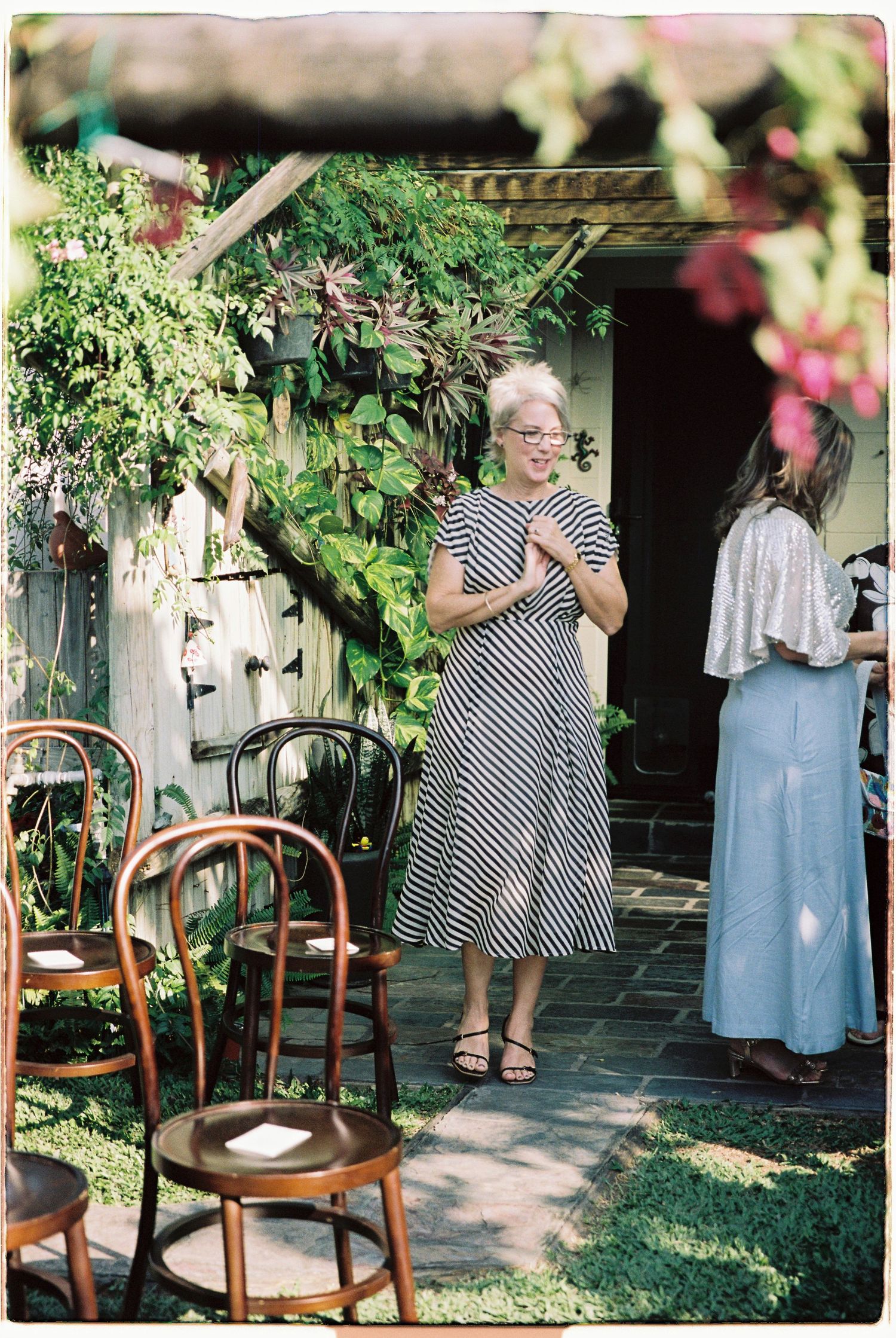 Two women in elegant dresses chatting outdoors near lush greenery and vintage wooden chairs at a garden event.