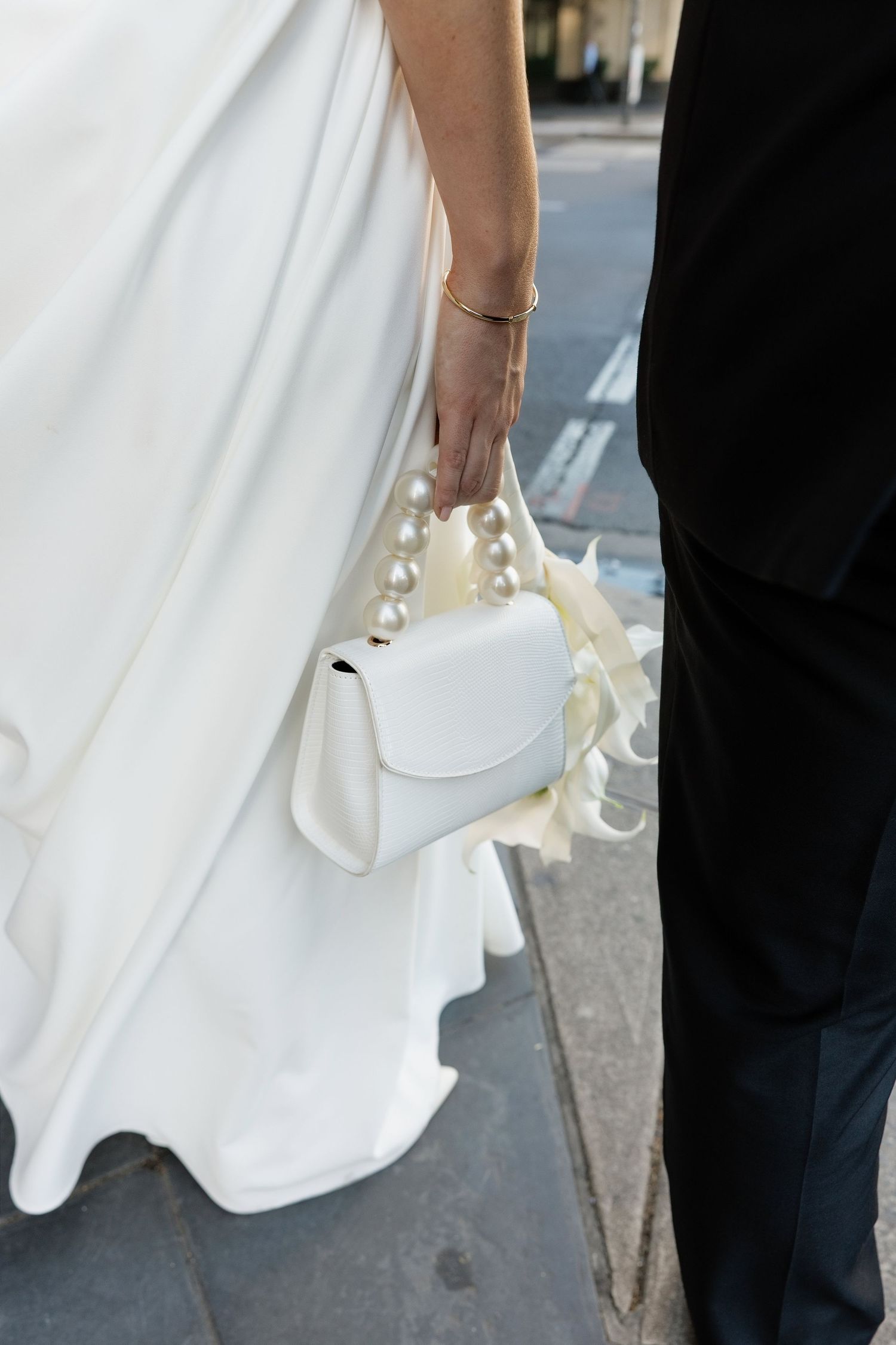 Close-up of bride holding white pearl-handled clutch bag, wearing white wedding gown and gold bracelet.