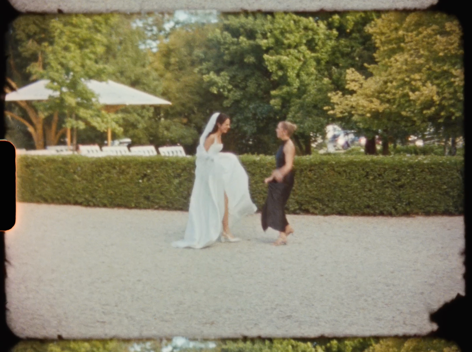Bride in a white gown and veil dancing playfully with a guest on a gravel path in a garden.