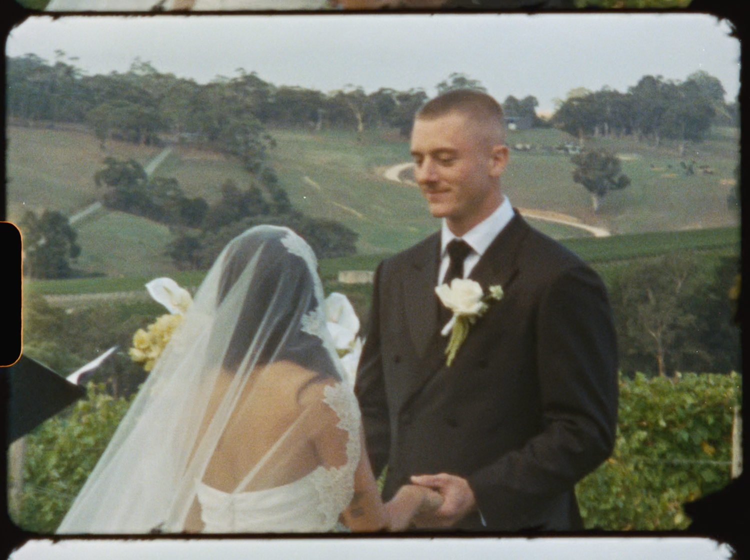 Groom smiling at bride during outdoor vineyard wedding ceremony with scenic hills backdrop.