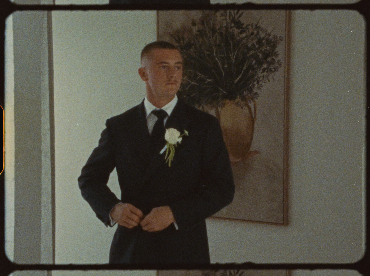 Groom in black suit with white rose boutonniere standing in front of floral painting.